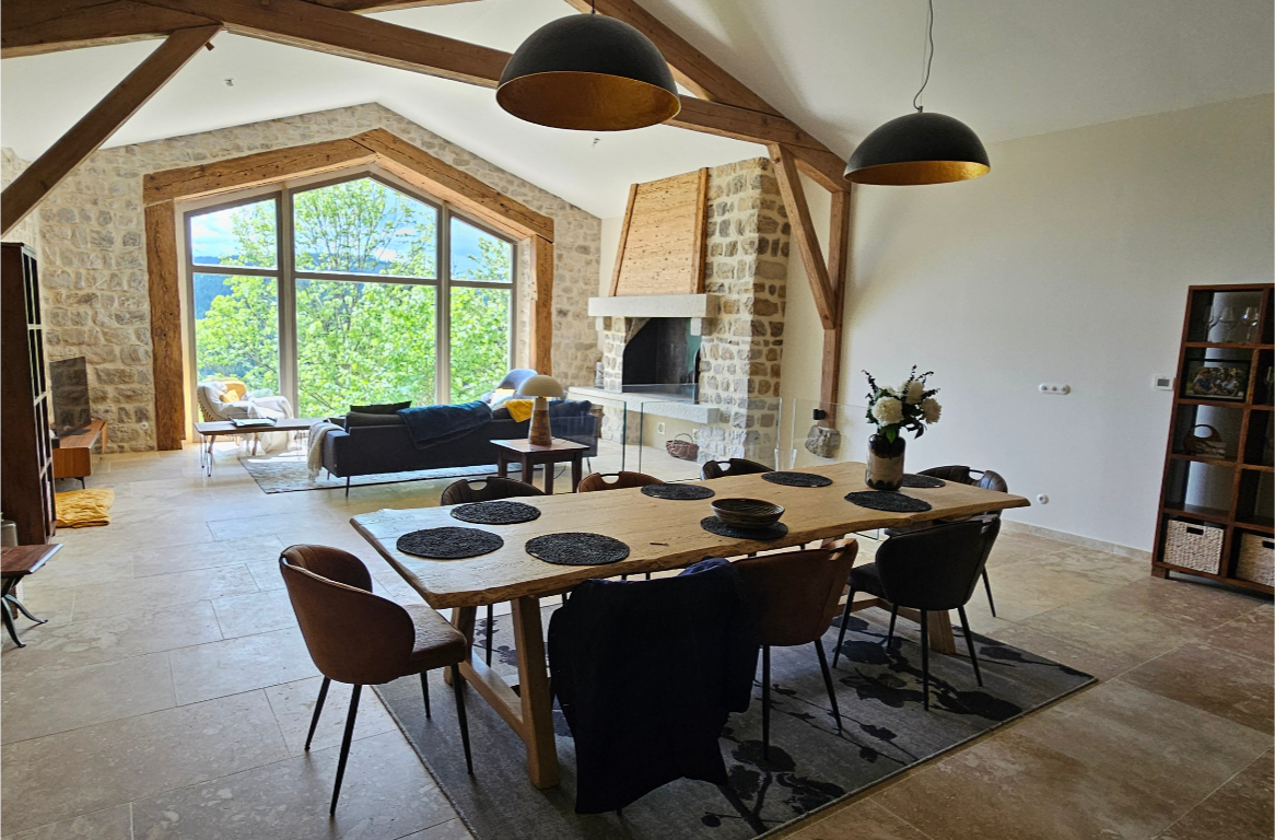 Salle à manger avec un grand table en bois, huit chaises, une cheminée en pierre, un grand mur en face avec de grandes fenêtres offrant une vue sur la nature, deux lampes suspendues noires, avec un salon au fond.