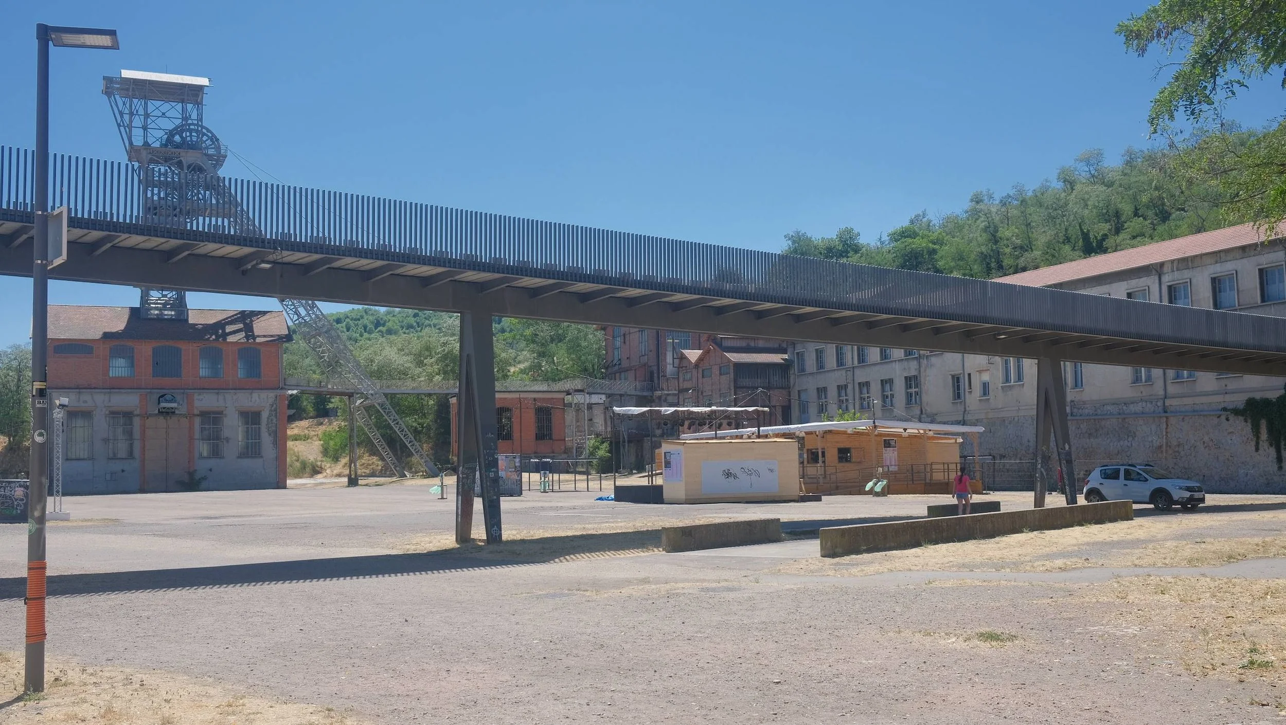 Vue d'une place urbaine avec un pont piéton, bâtiments industriels, arbres en arrière-plan et un ciel bleu.