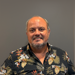 Homme avec barbe portant une chemise à motifs floraux, souriant devant un mur gris