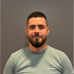 Portrait d'un homme avec une barbe, portant un t-shirt gris clair, devant un mur gris.