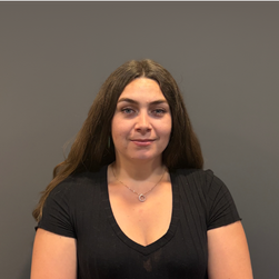 Jeune femme avec longs cheveux bruns, vêtue d'un t-shirt noir, souriant légèrement, contre un fond gris.