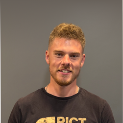 Jeune homme avec des cheveux blonds et une barbe, portant un t-shirt noir, souriant contre un mur gris.