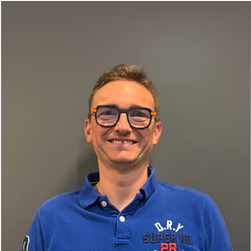 Jeune homme souriant portant des lunettes et un polo bleu, debout devant un mur gris.