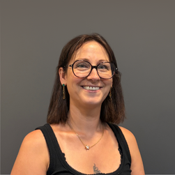 Portrait d'une femme souriante portant des lunettes et un haut noir, contre un mur gris.