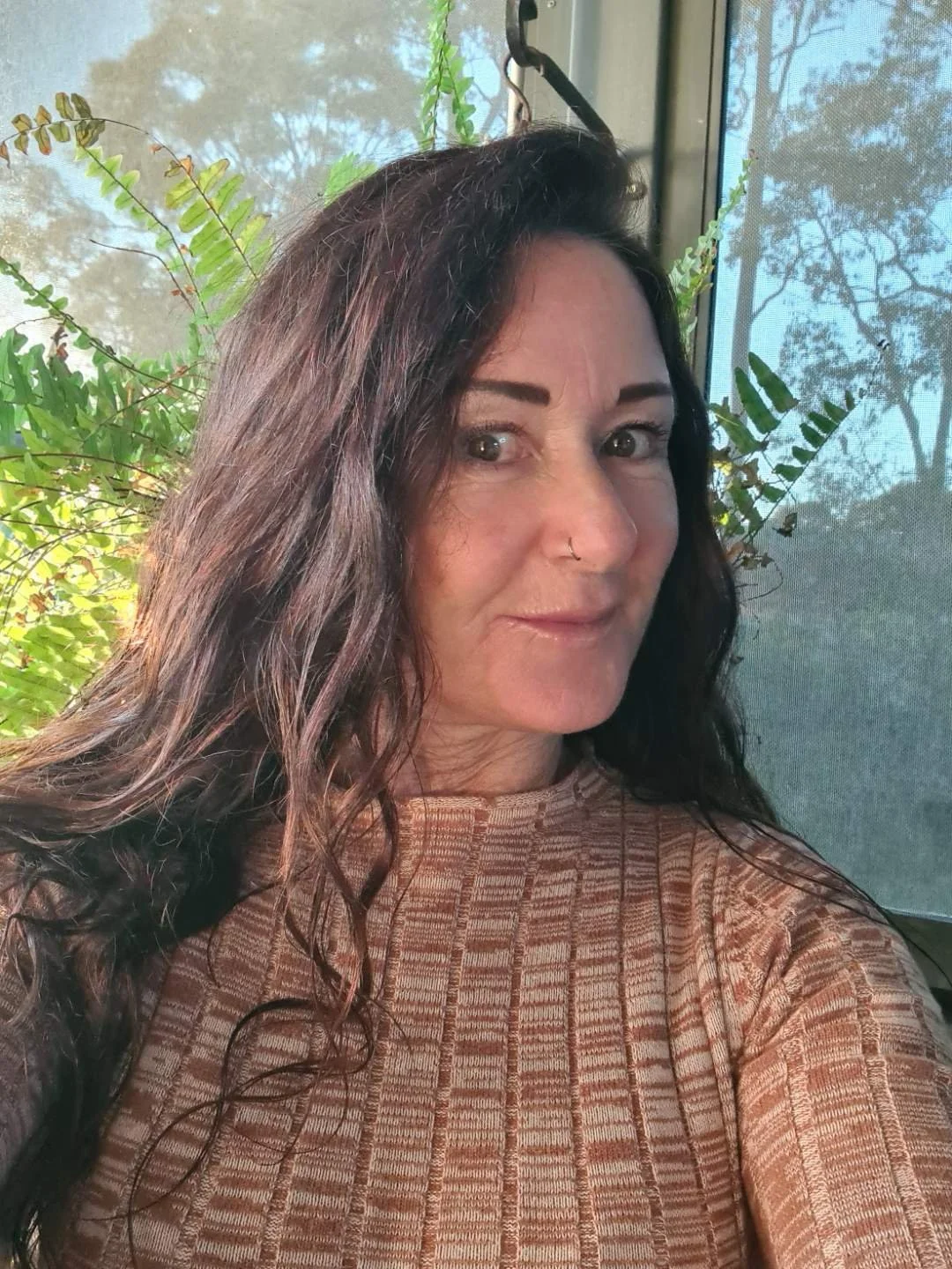 A woman with long curly dark hair, light skin, and a nose piercing taking a selfie indoors near a window with greenery outside.