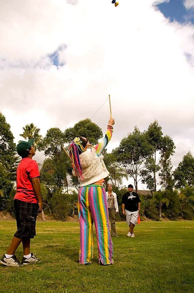 A woman with rainbow-colored striped pants and multicolored hair takes a selfie with a selfie stick while three children and adults watch on in a park with grass and trees.