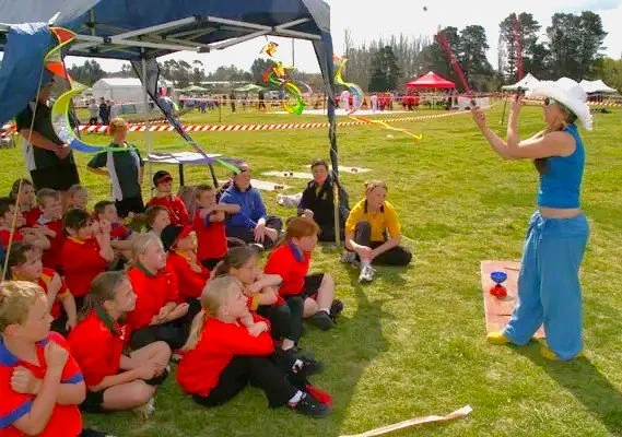 A group of children sitting on the grass watching a woman in a blue outfit and white hat perform a juggling act at an outdoor event.