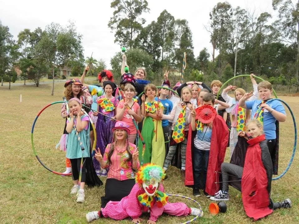 Children dressed as clowns and circus performers holding hoops and props outdoors on a grassy field.