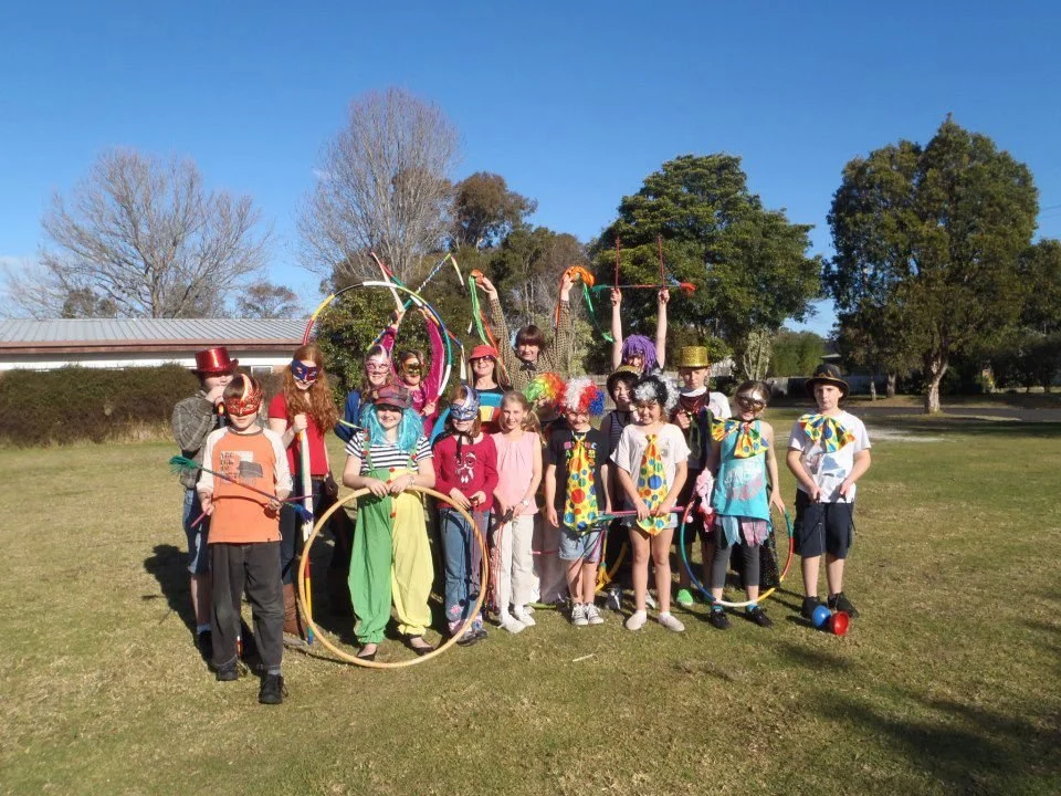 Children dressed as clowns with colorful costumes, balloons, and props outdoors on a sunny day.