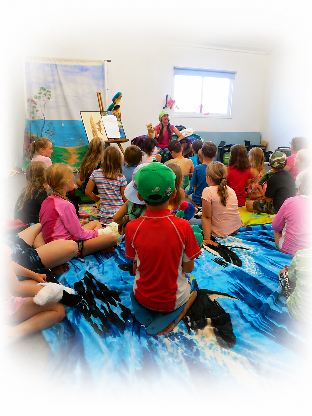 Children sitting on a colorful blanket, watching a puppet show in a bright room with a painted backdrop and a window.