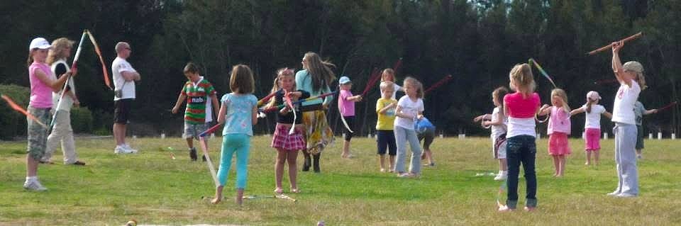 Children playing with sticks or staffs in an open grassy park on a sunny day.