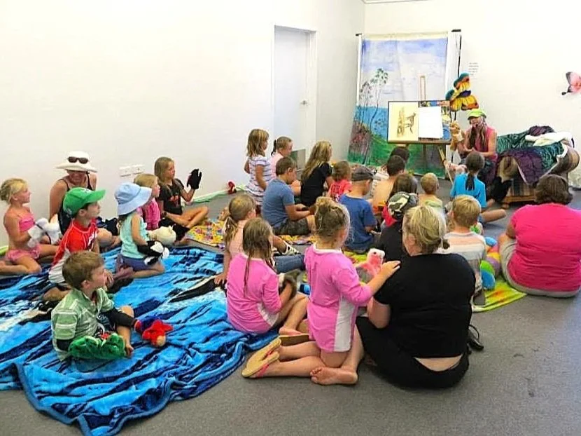 Children and adults sitting on blankets in a room, watching a storytime presentation by a woman dressed as a clown with a puppet.