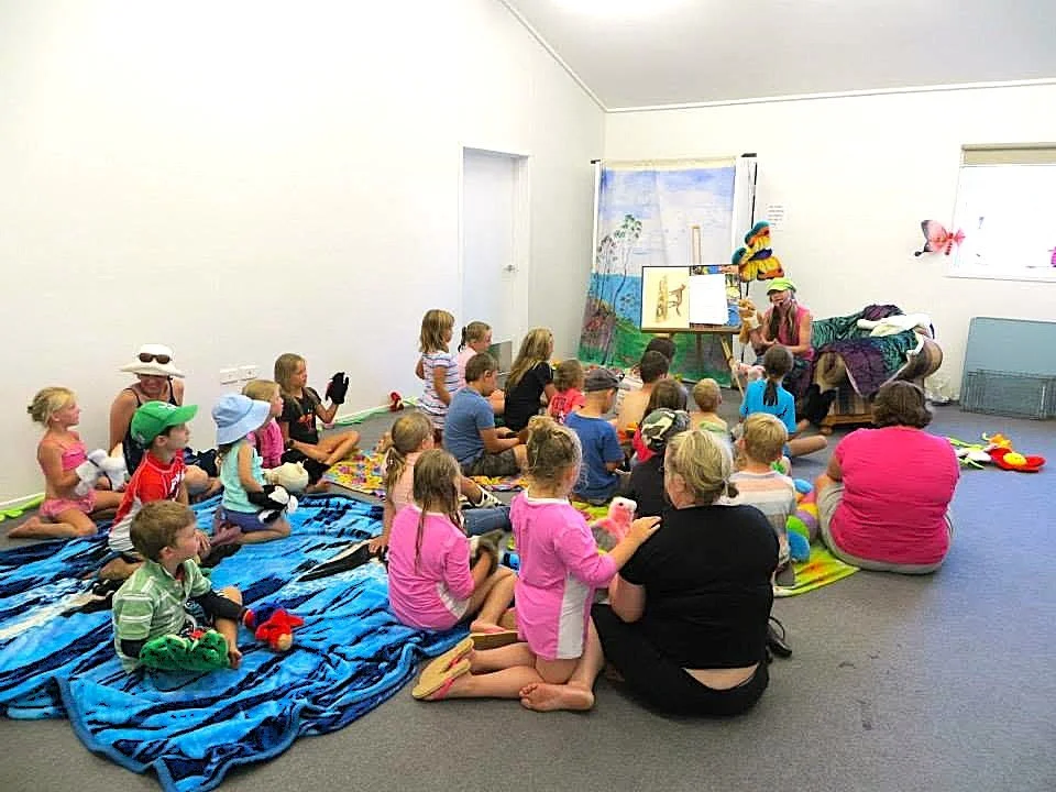 Children sitting on colorful blankets and mats, watching a woman perform puppet shows or storytelling in a room decorated with beach and butterfly-themed artwork.