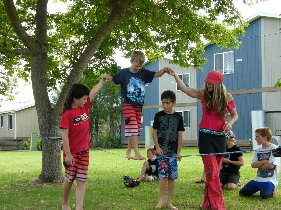 Children and adults at a park engaging in a tightrope walk activity between two trees.