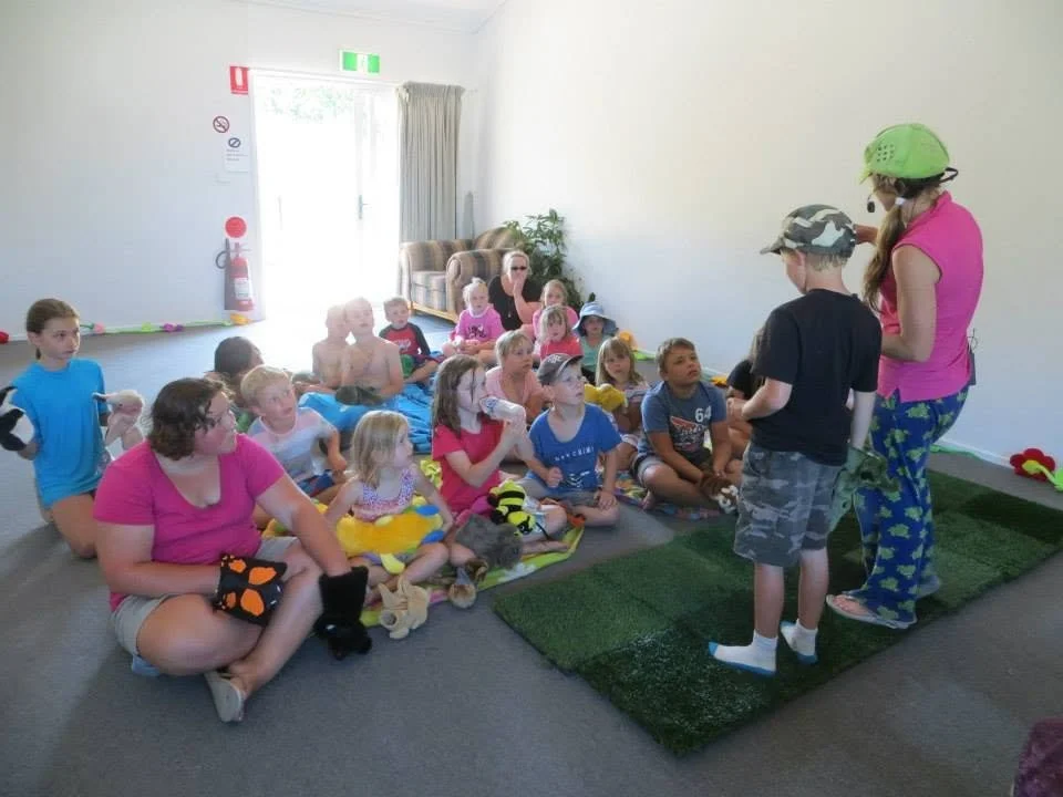 Children gathered indoors sitting on a blanket, listening to a woman and a boy standing in front of them, engaging in a storytelling or activity.