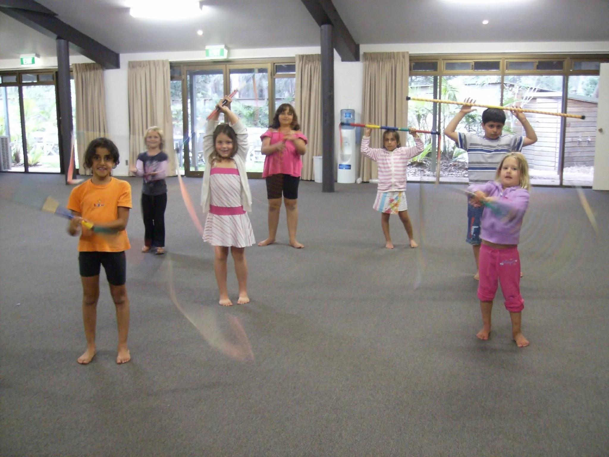 Kids posing with sticks indoors, some children holding sticks above their heads, others holding sticks at different angles, standing barefoot on a carpeted floor, with large windows and a water cooler in the background.