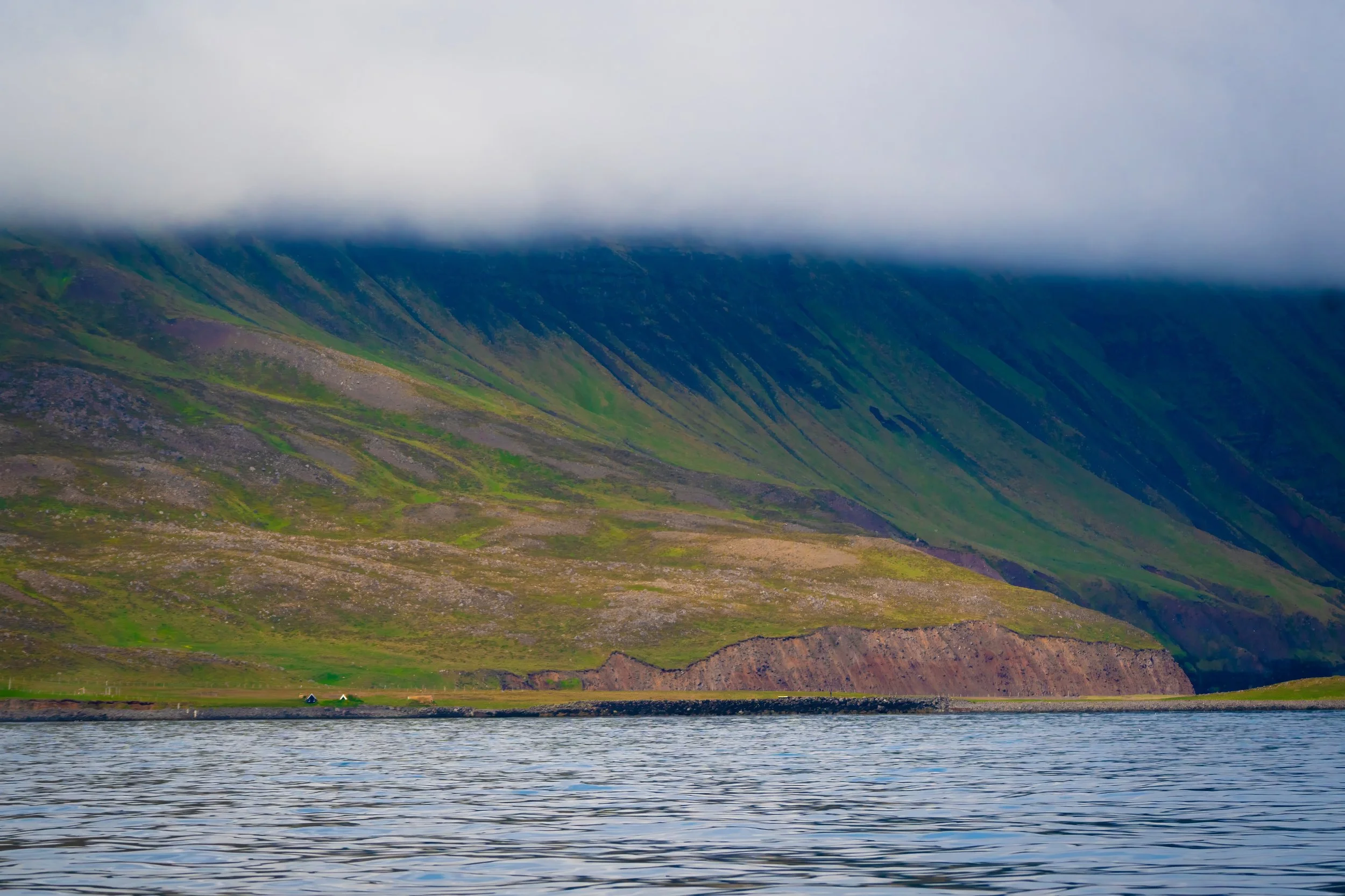 Peaceful lake with gentle ripples in the foreground, lush green hillside with purple and brown patches rising behind, topped with thick gray clouds partially obscuring the mountain peaks.