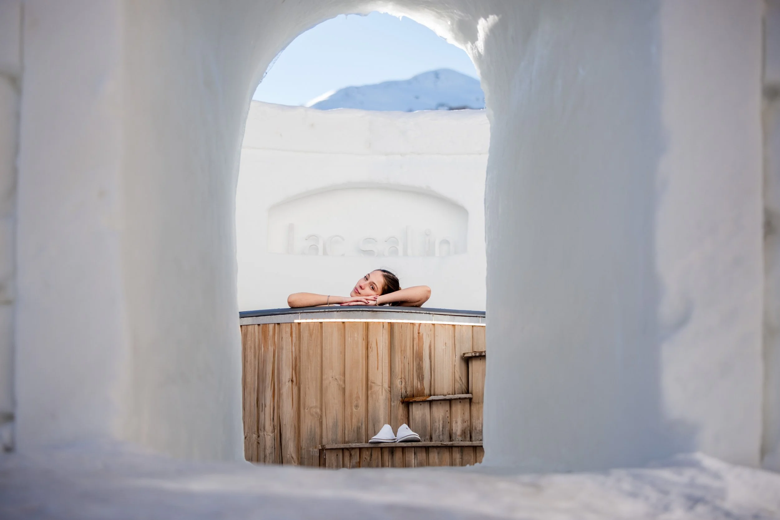 A young woman resting her head on her arms, lying on a wooden platform, seen through a snow tunnel with a mountain in the background.