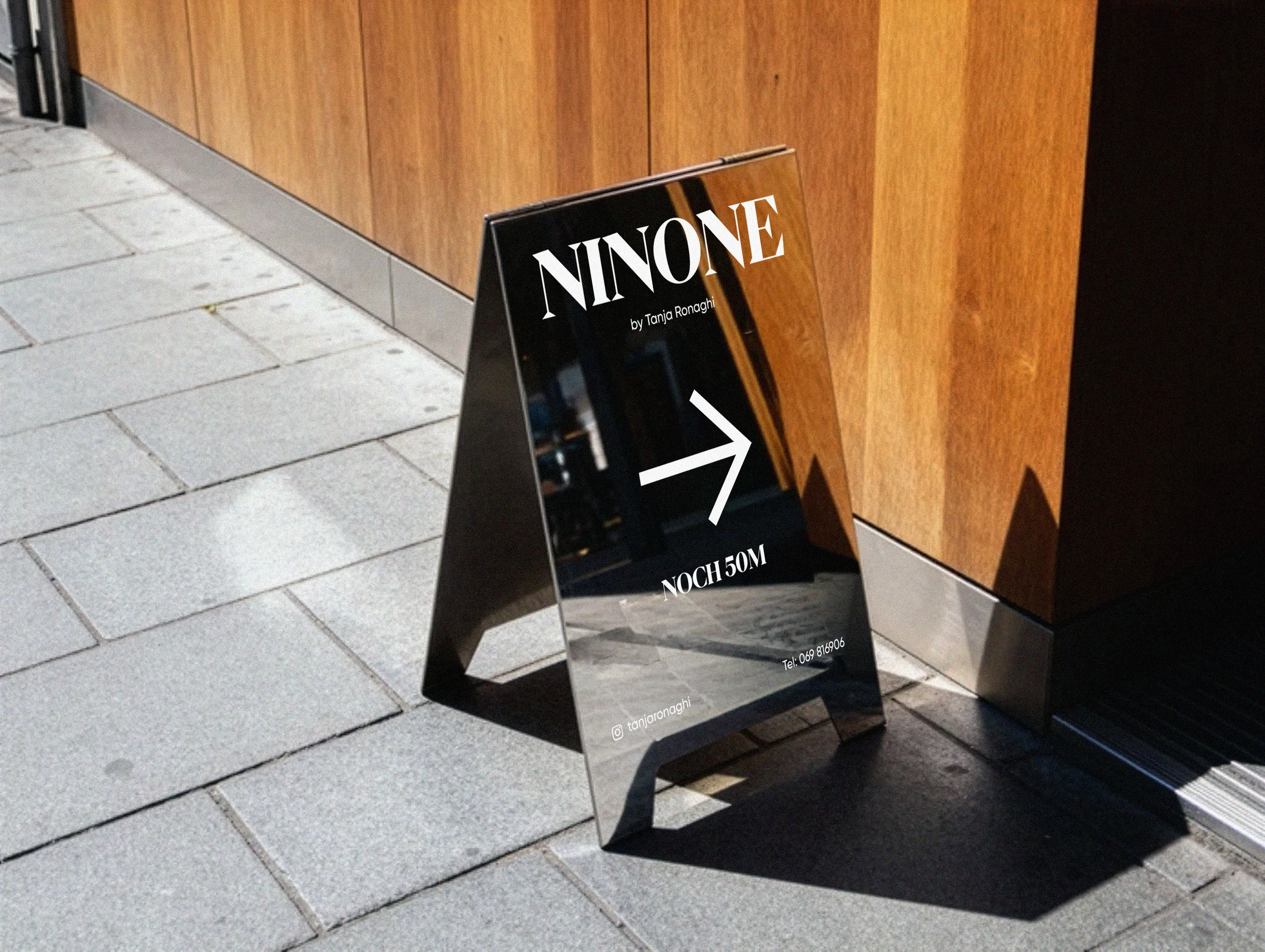 Ein schwarzer Werbeschreibtisch mit weißen Buchstaben, der 'NINONE', 'by Tanja Ronaghi' und Kontaktinformationen zeigt. Der Tisch steht auf einem Gehweg vor einer Holzfassade.