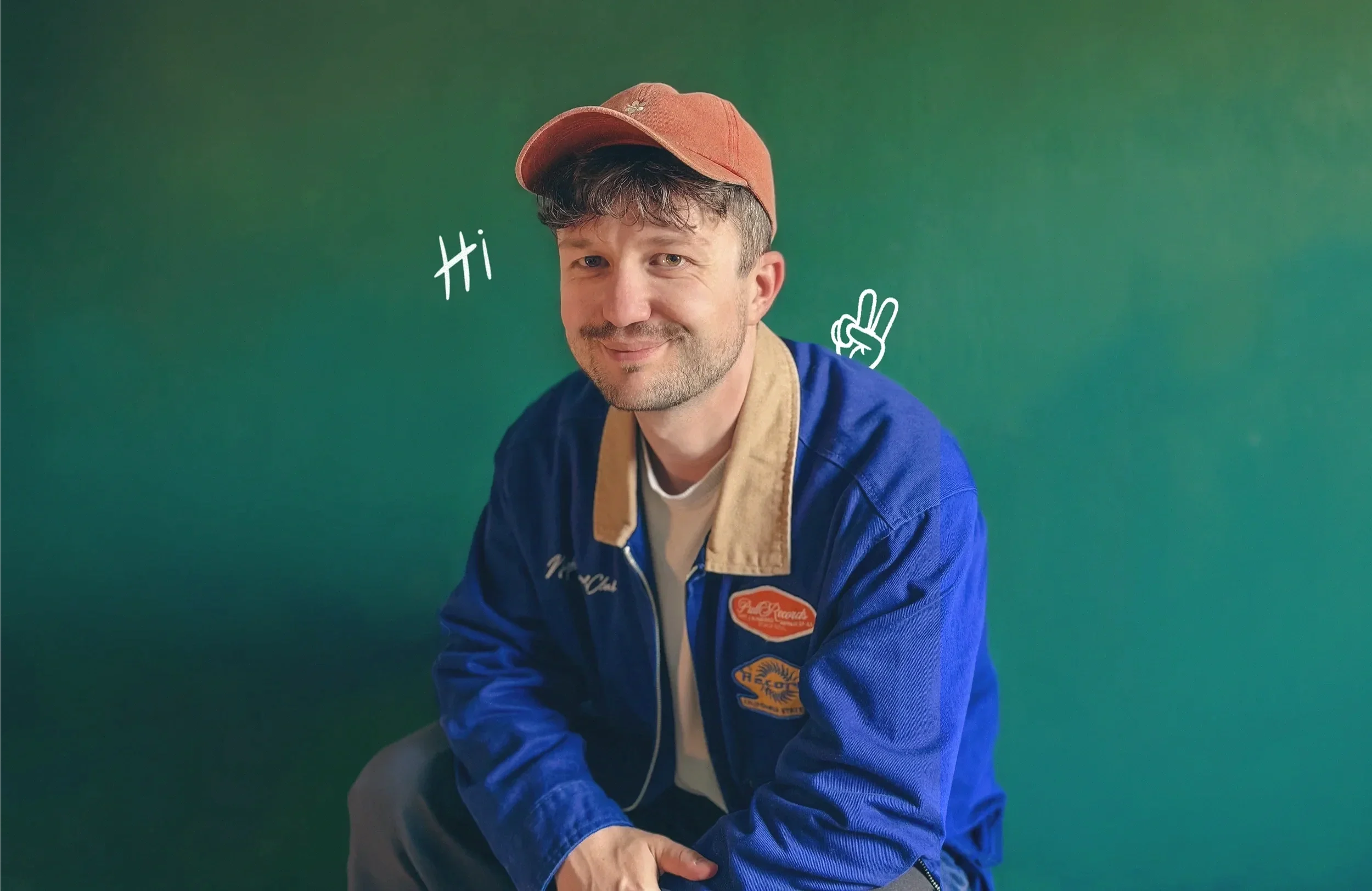 Ein jüngerer Mann mit braunem Haar, blauen Augen und leichtem Bart trägt eine braune Kappe und eine blaue Jacke mit aufgenähten Patchs. Er lächelt leicht und sitzt vor einem grünen Hintergrund, wobei er nach rechts schaut. Text und Handzeichnung nebeneinander, die auf seine Schulter zeigen, sagen "Hi" und zeigen eine Hand, die ein Peace-Zeichen macht.