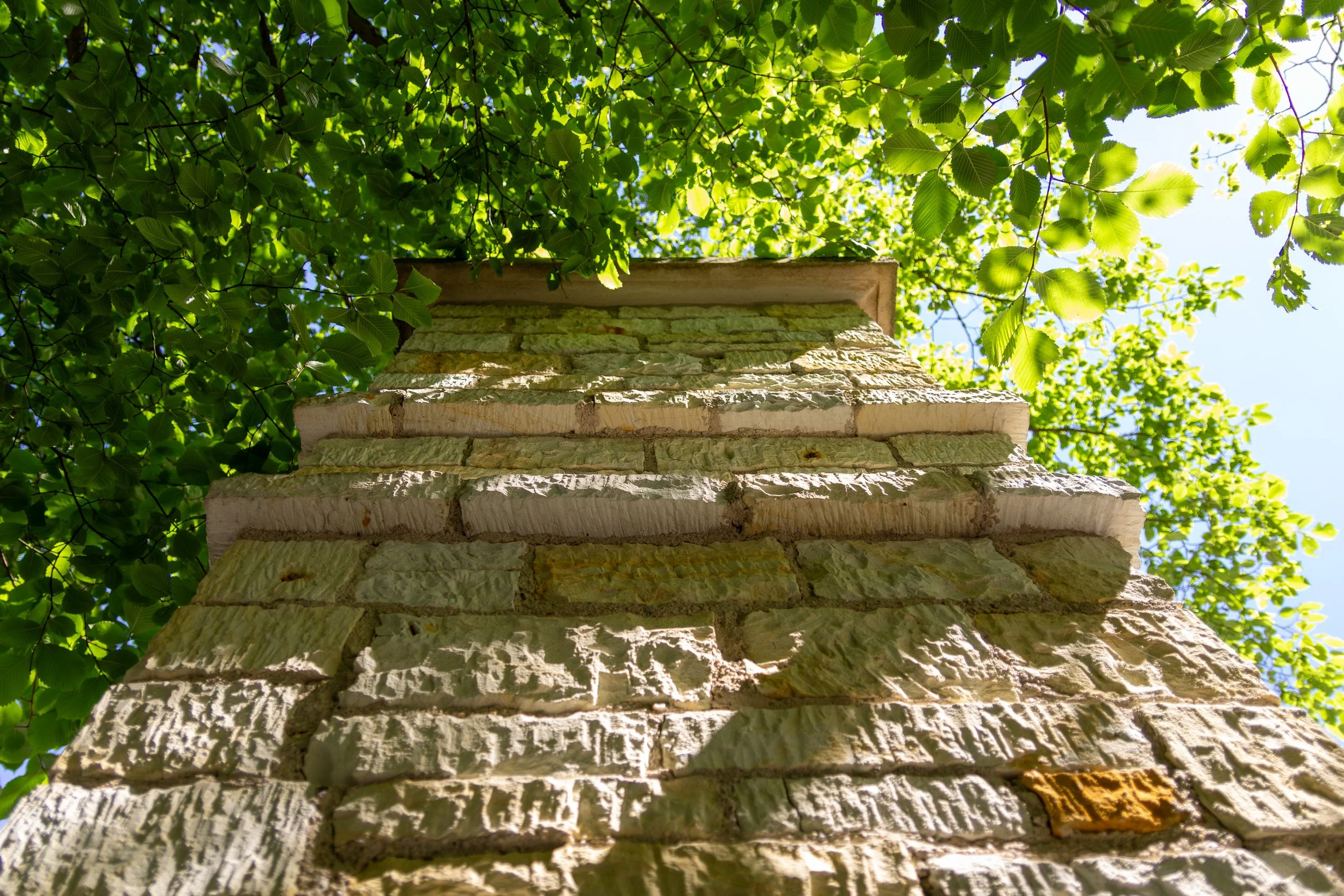 Low-angle view of a stone column with green leafy tree canopy overhead and patches of blue sky visible.
