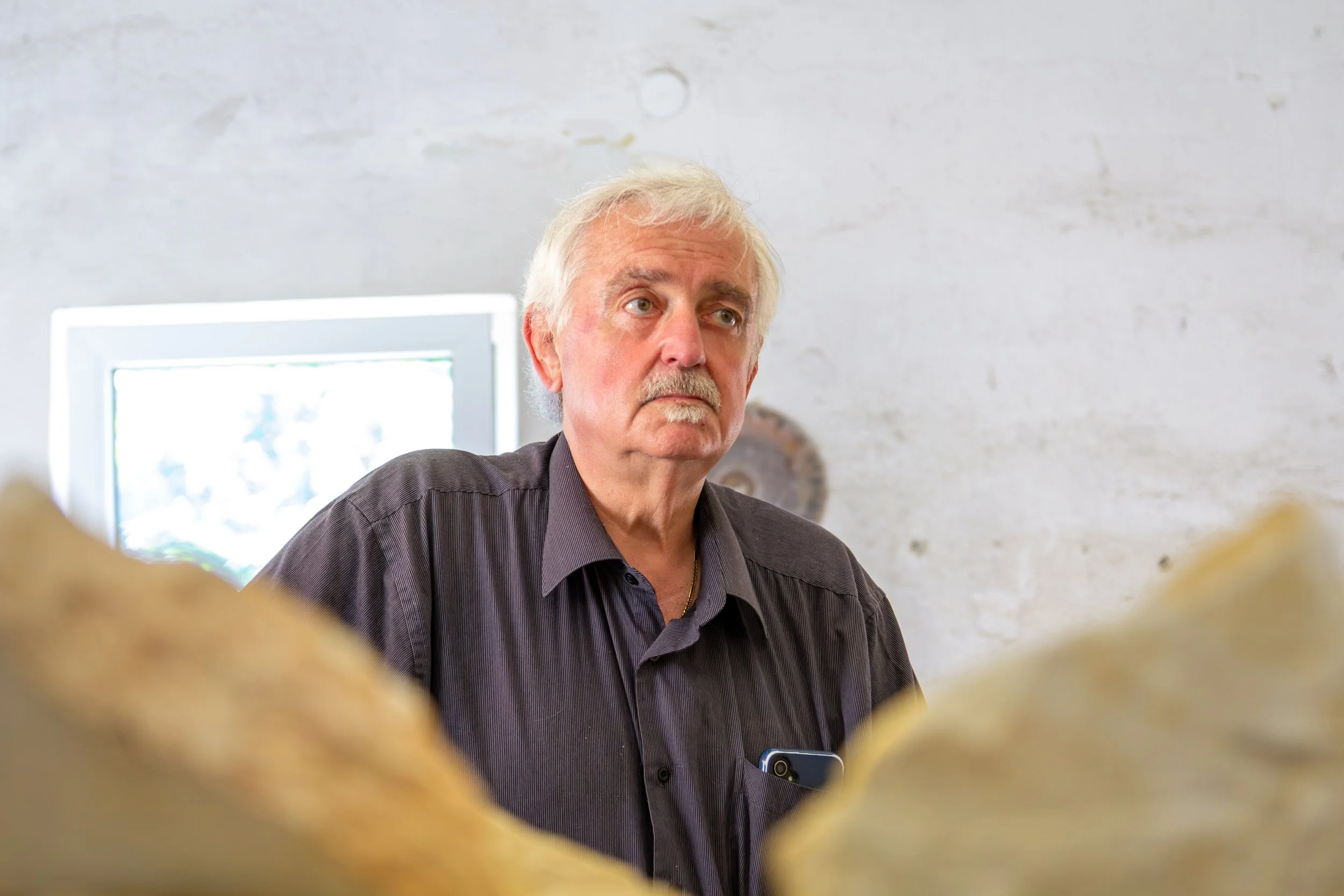 An older man with white hair and a mustache, wearing a dark shirt, sitting indoors with a white wall and a window behind him, looking to his left with a serious expression.