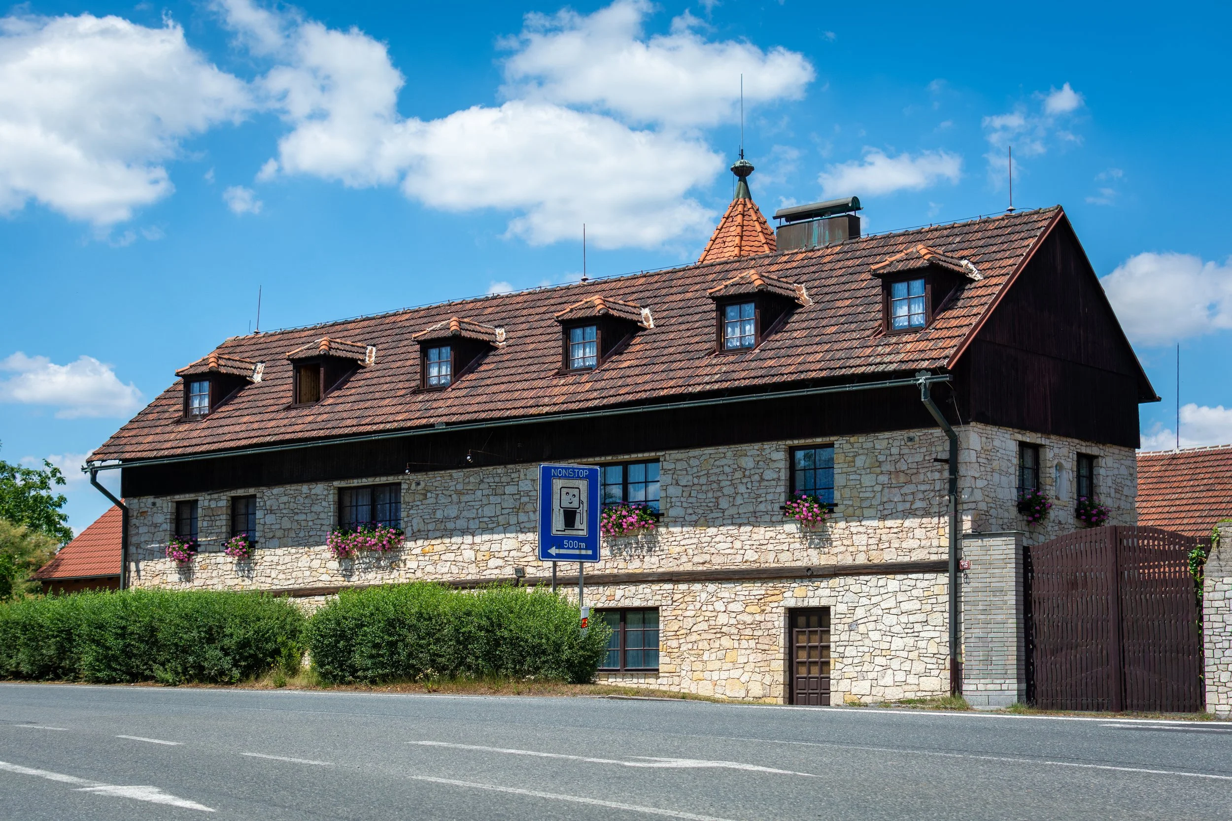 A large building with a stone facade, multiple dormer windows, and a steep roof with red tiles. There are colorful flowers hanging outside the windows and a street sign in the foreground, with a mostly clear blue sky and a few white clouds overhead.