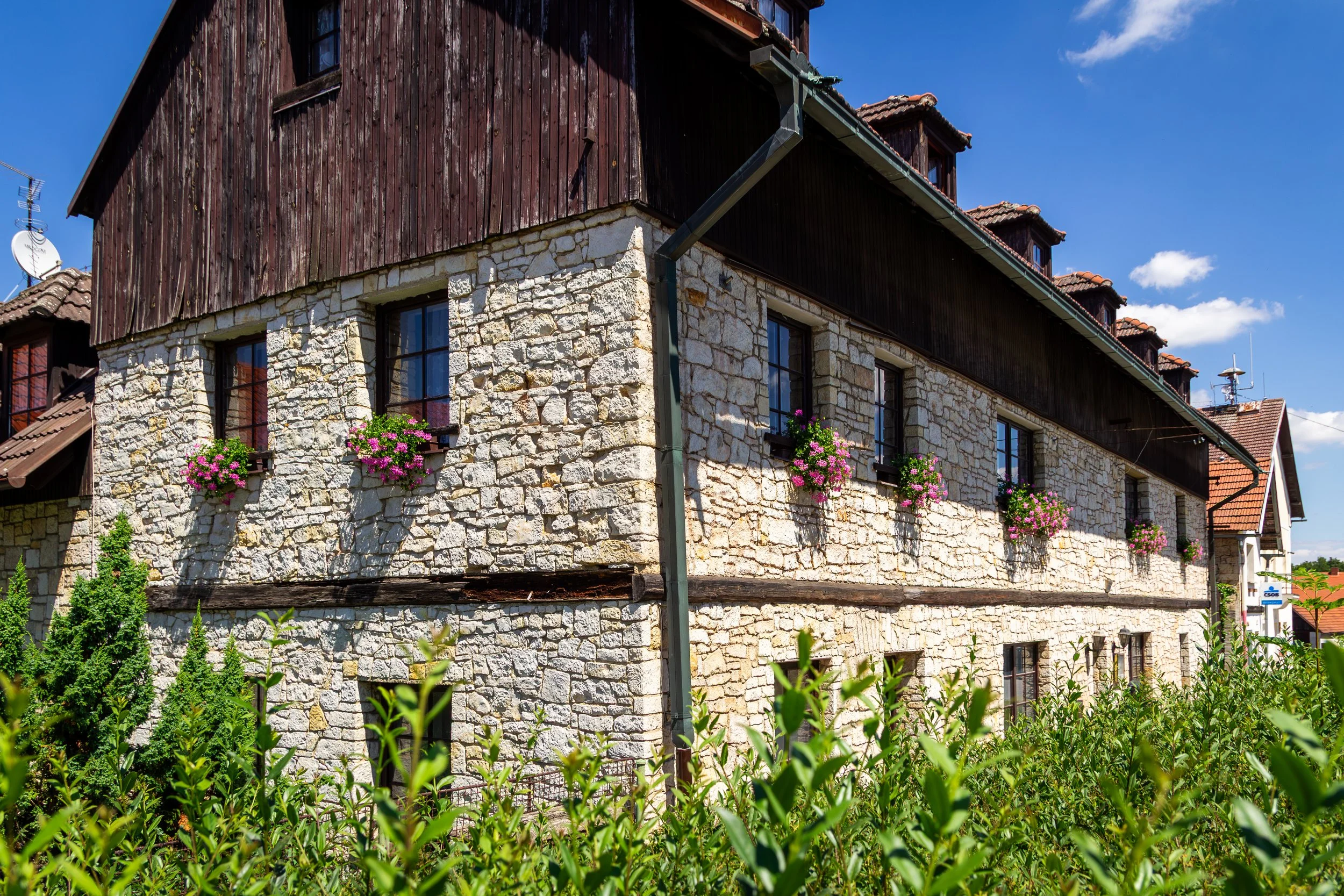 A multi-story building with a stone lower level and a wooden upper level, featuring mounted flower baskets with pink flowers, under a bright blue sky with some clouds.