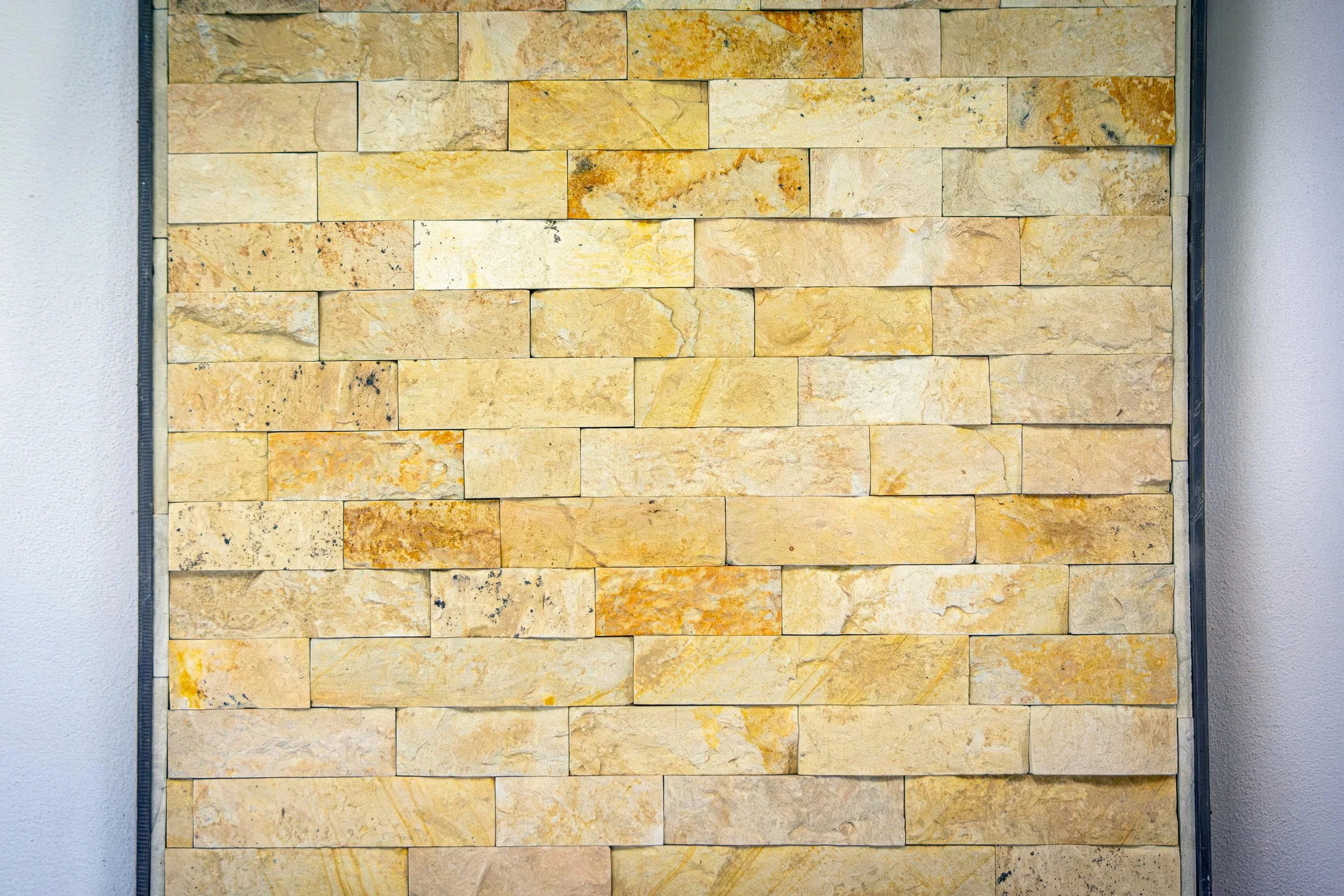 A wall made of rectangular beige and tan stones with various textures and patterns, framed by a narrow metal boundary.