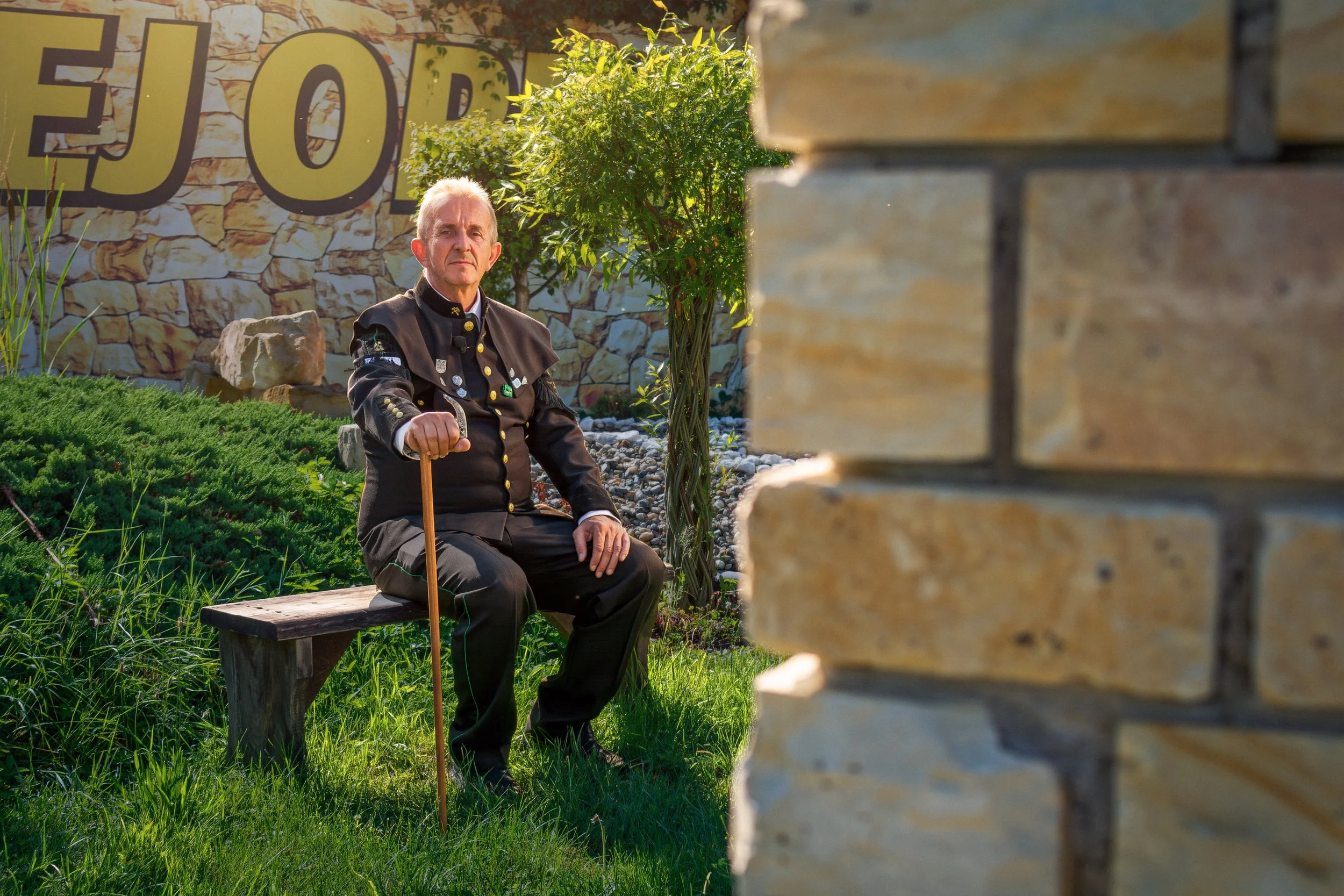 A man dressed in traditional Bavarian clothing sitting on a wooden bench outdoors, holding a walking stick, with trees and a large yellow sign with black letters in the background. The scene is lit by sunlight and includes greenery and decorative rocks.