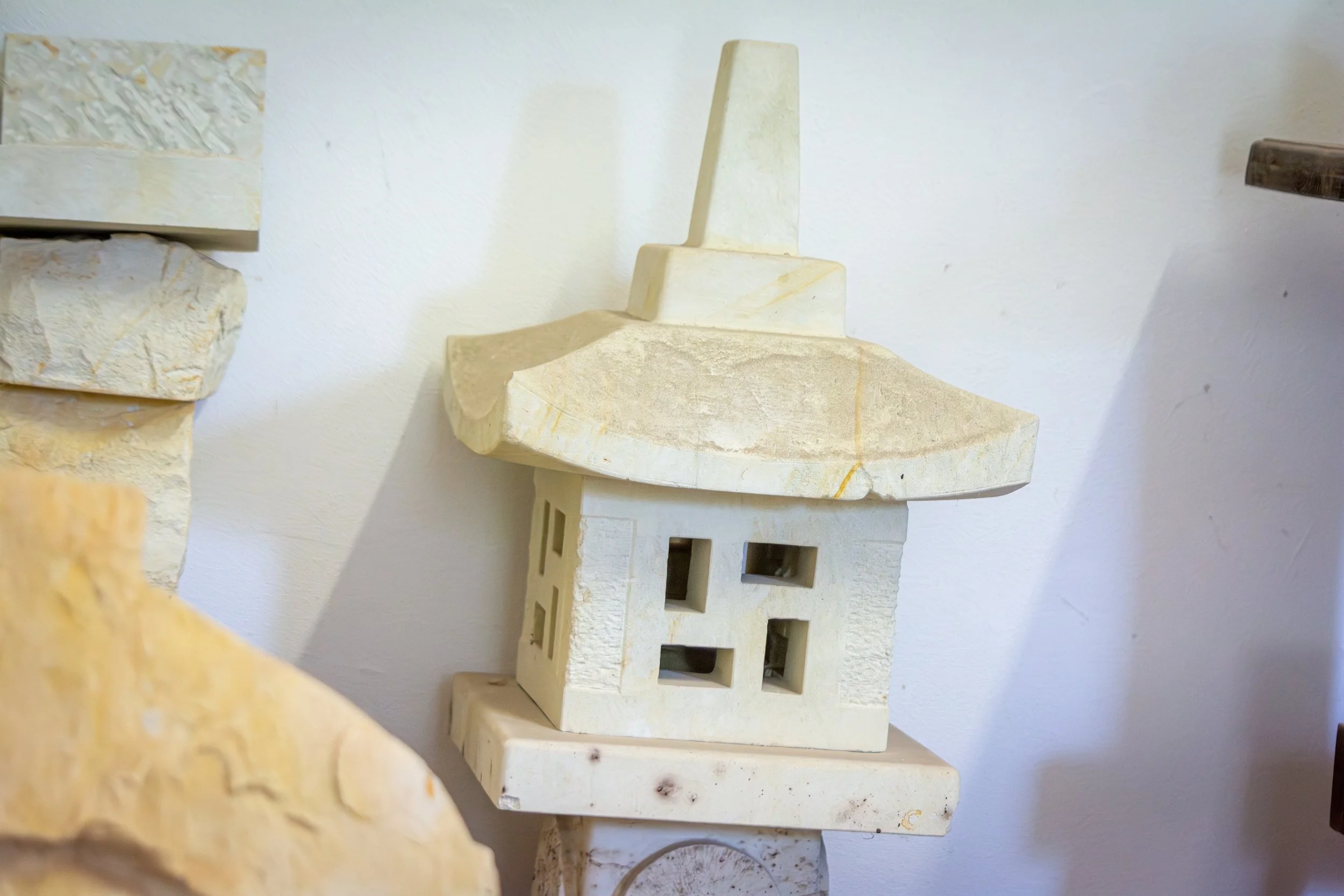 Close-up of beige stone architectural model with a building featuring multiple windows and a sloped roof, against a white wall background.