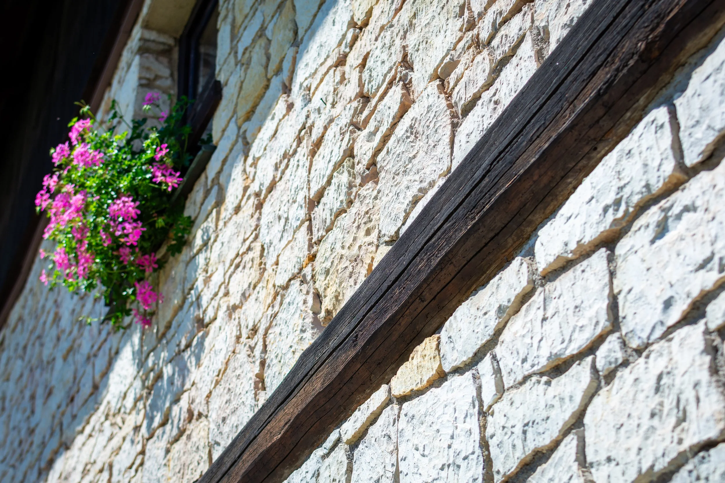 Close-up of a white brick wall with a dark wooden beam running diagonally across it and a window with pink flowers in a planter box.
