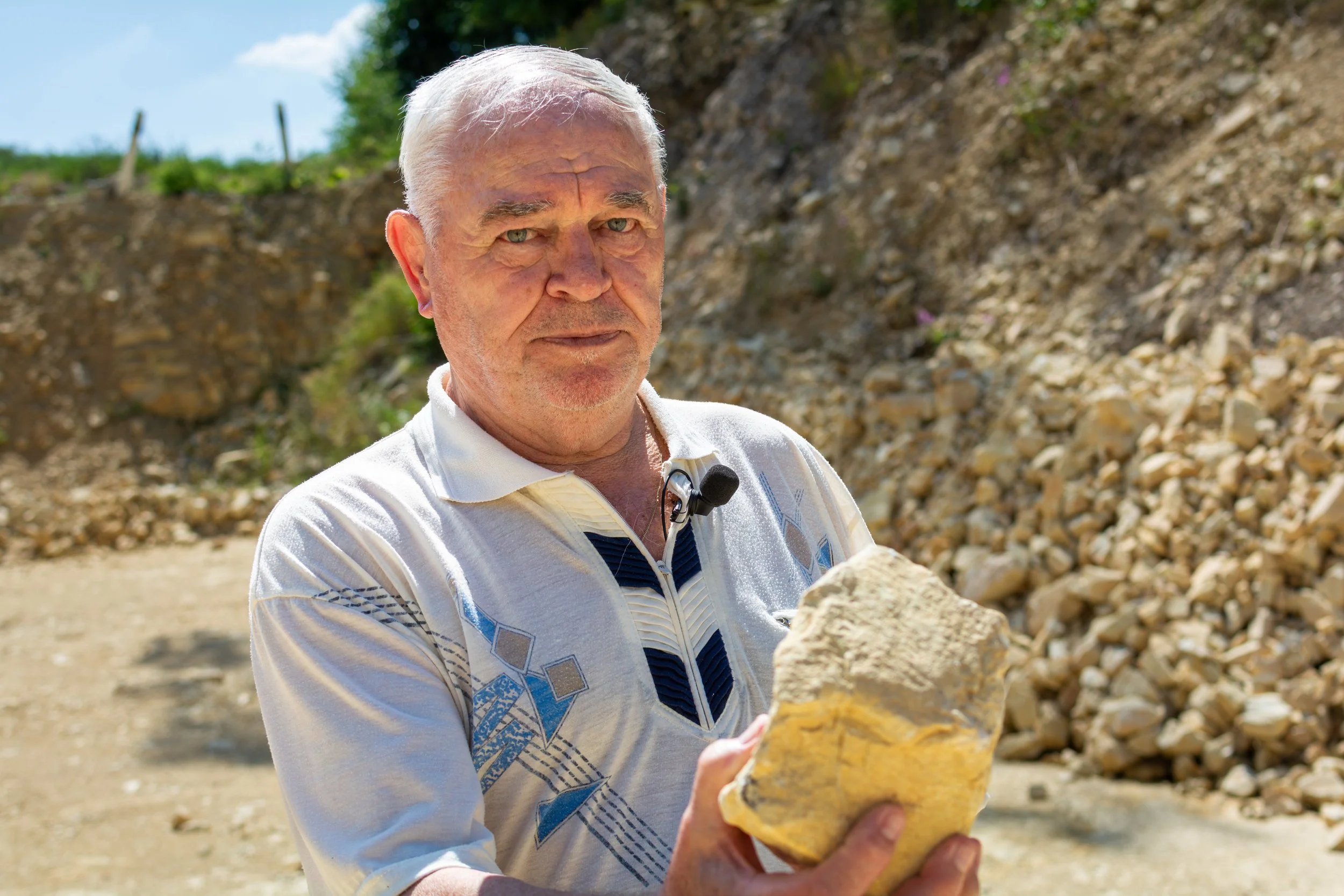 An elderly man with gray hair and a white polo shirt outdoors holding a large yellowish rock.