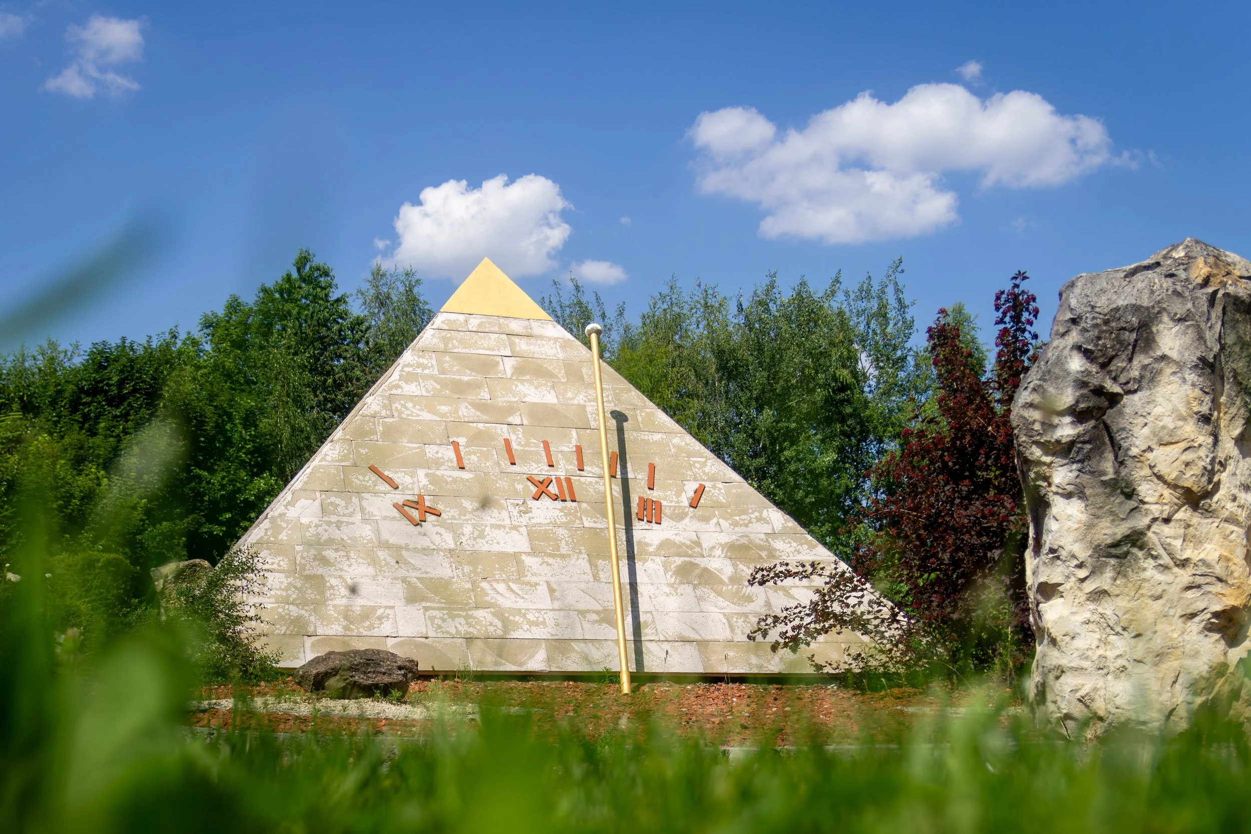 A stone outdoor clock with Roman numerals and a triangular top, surrounded by green trees and rocks, under a blue sky with clouds.