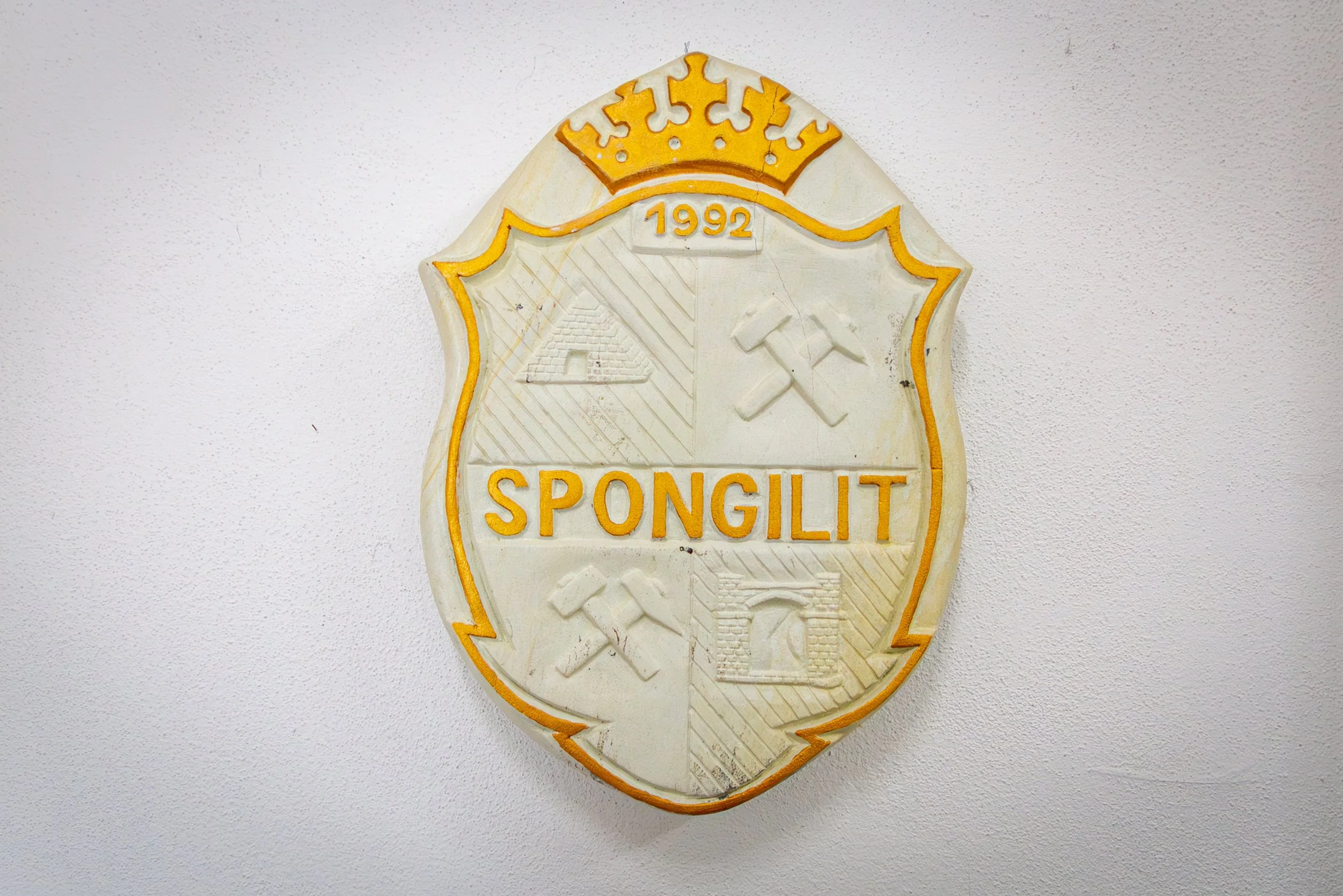 A cream-colored emblem with yellow outlines and features a crown at the top, the year 1992 in the center, and the words 'Spongillit' at the bottom, mounted on a white wall.