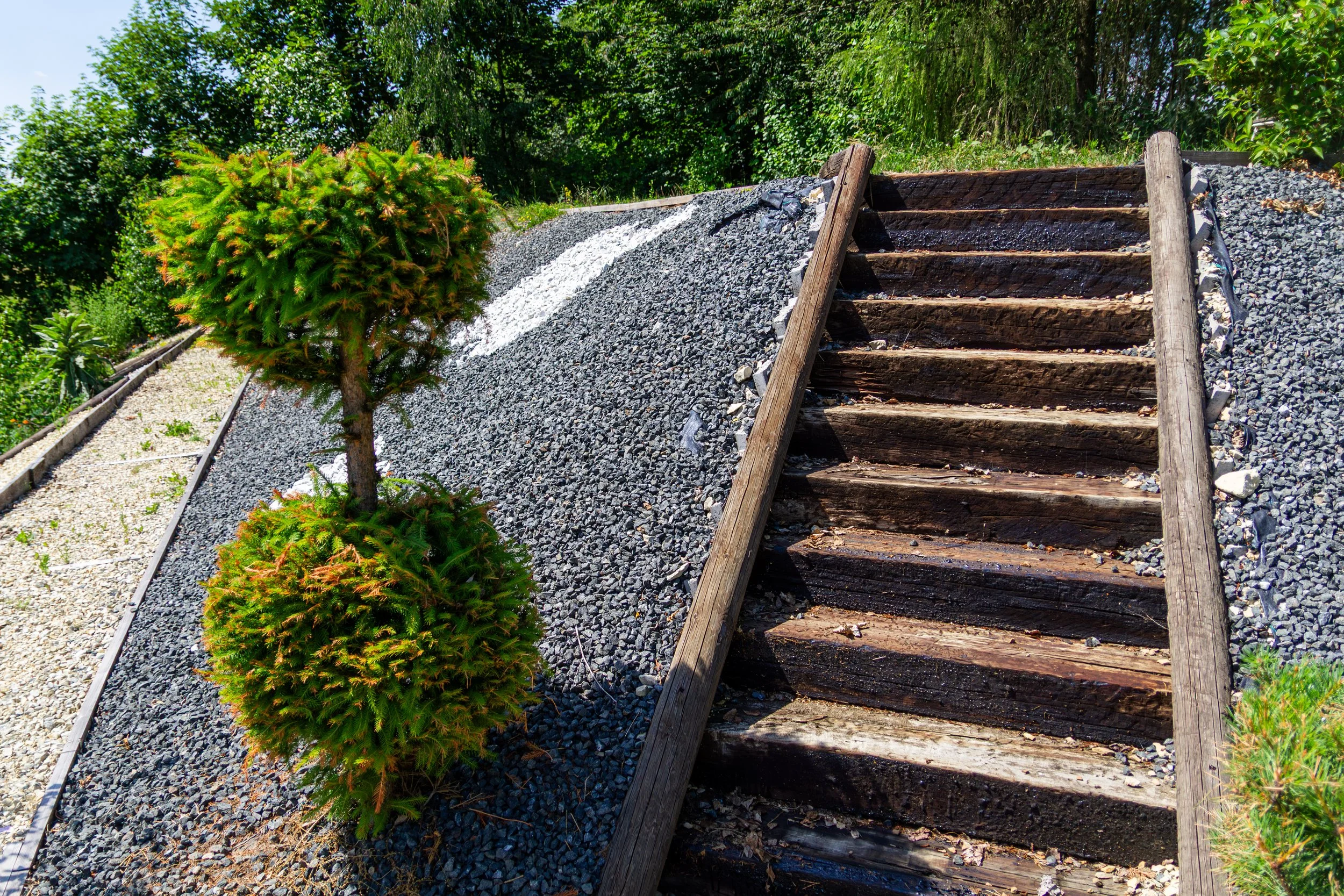 A set of outdoor wooden stairs with gravel on the sides and a small green bush in front. The staircase ascends beside a gravel incline and is surrounded by green trees and bushes under a blue sky.