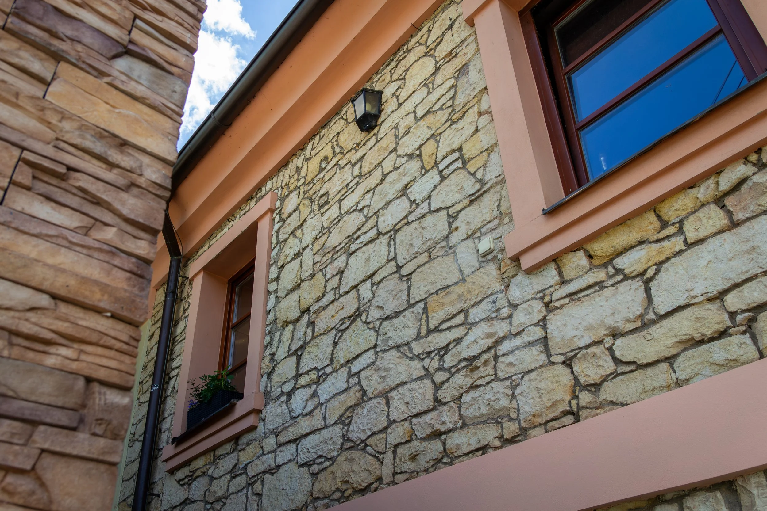 Exterior of a house showing a stone wall, a window with a brown frame, a black wall-mounted light fixture, and a corner with a brick wall at an upward angle under a partly cloudy sky.