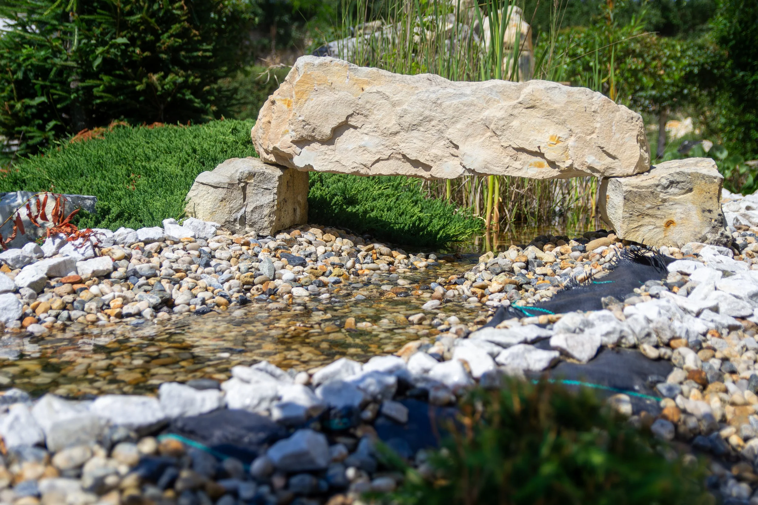 A small landscaped garden with a stone bridge over a shallow stream, surrounded by green plants and rocks.