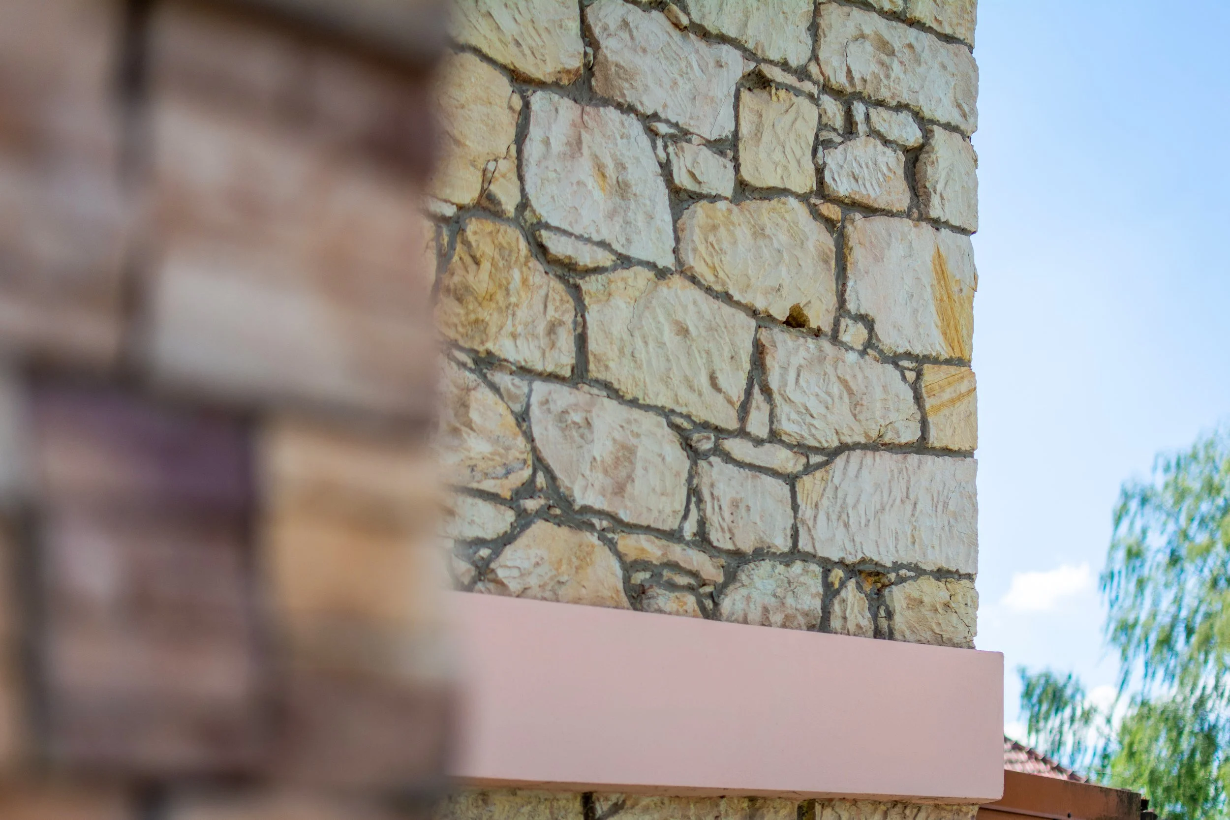 Close-up of a textured stone wall with light-colored stones and dark grout, with a portion of blurred brick wall and blue sky in the background.
