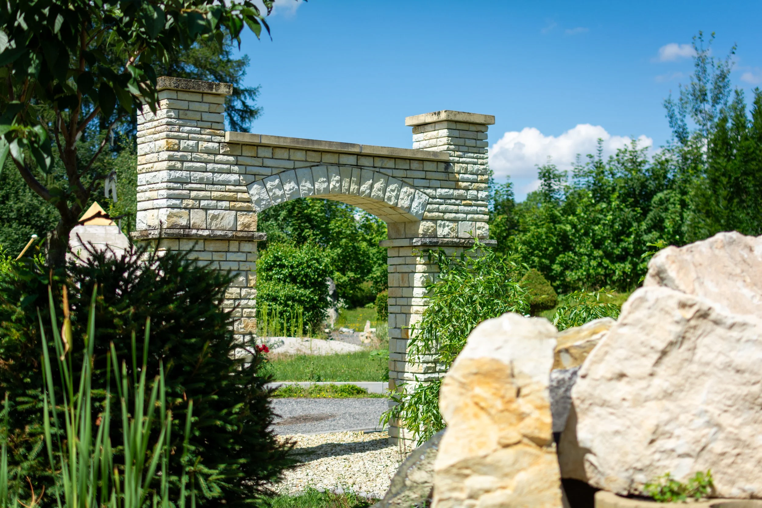 A garden scene with a stone archway supported by two stone pillars, surrounded by lush green trees and plants, under a blue sky with scattered clouds.