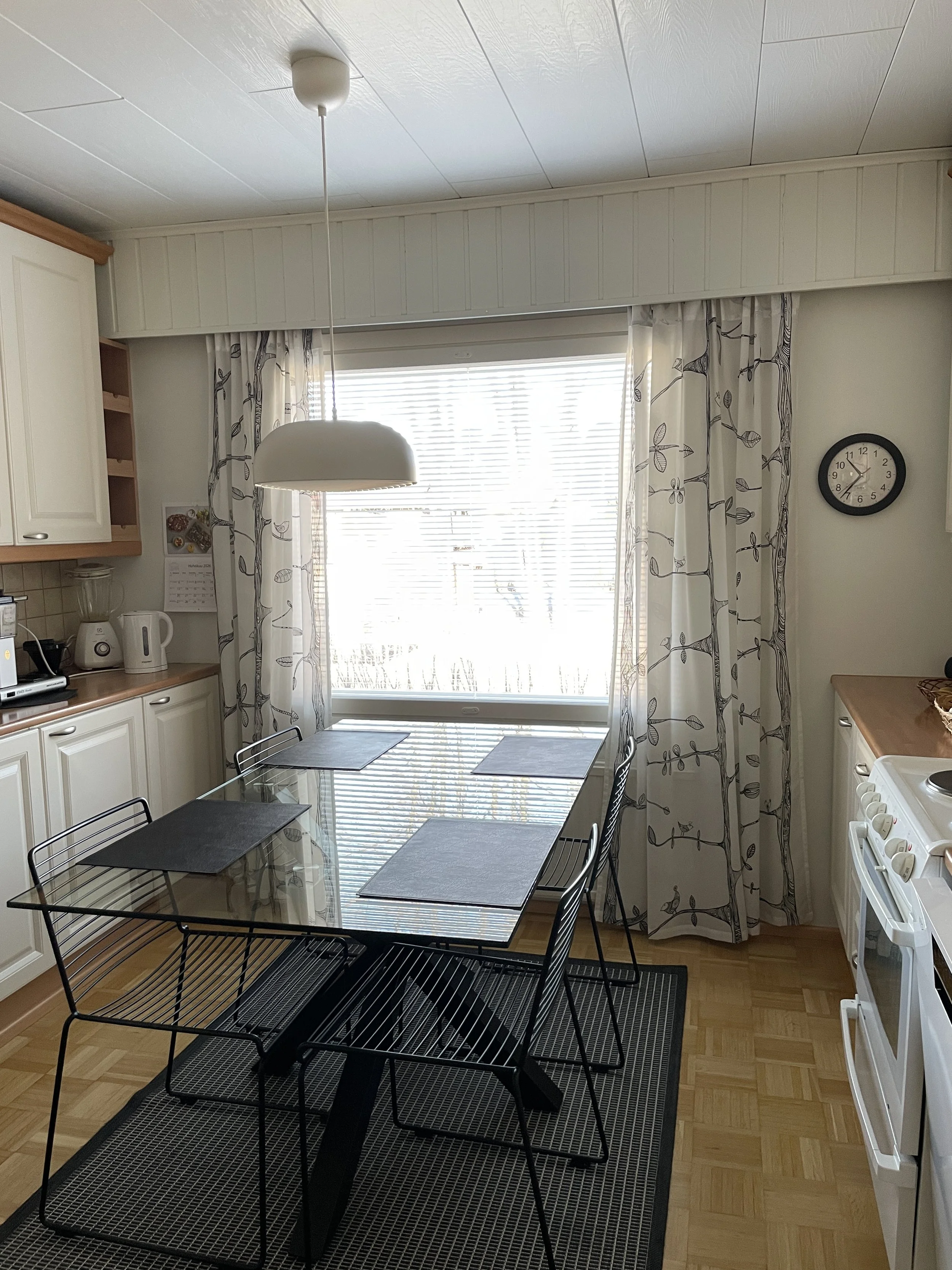 Keittiö, jossa on ruokapöytä ja neljä metallituolia, vaaleansävyiset verhot ja ikkunan edessä valkoinen lamppu.