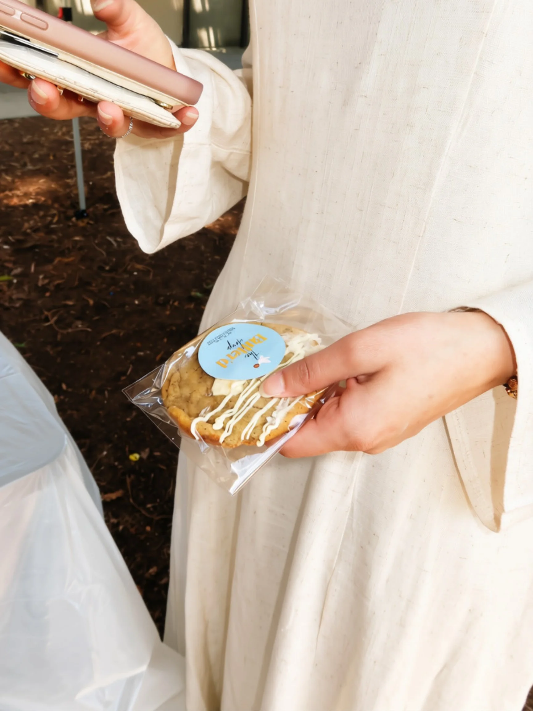 Person holding a packaged cookie with white drizzle, standing near a beige wall with a white cloth hanging on it, and an outdoor dirt ground in the background.