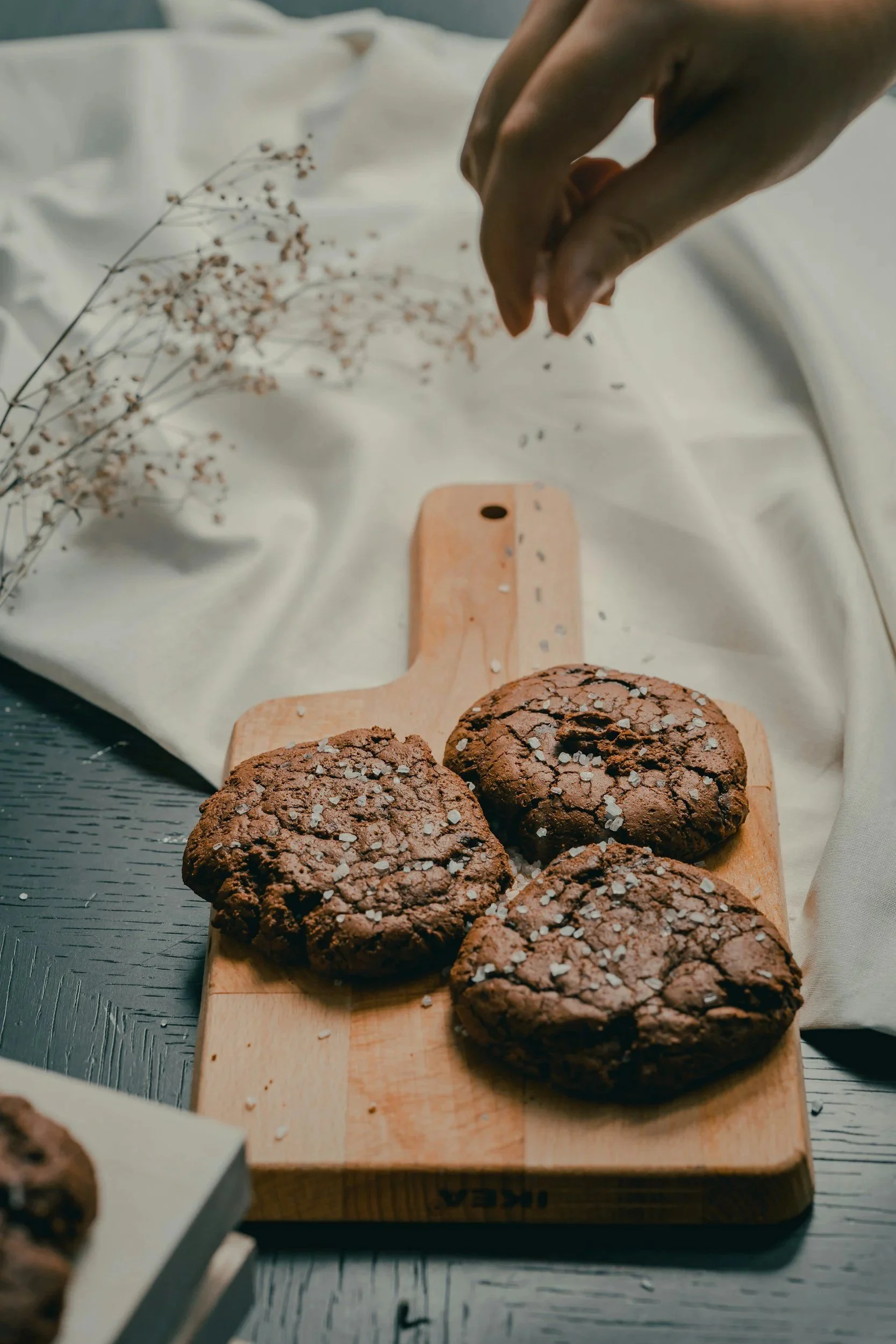 Close-up of a hand sprinkling salt onto a row of cookies with a textured surface, placed on a wooden cutting board. In the background, there is a sprig of dried flowers and a white cloth.