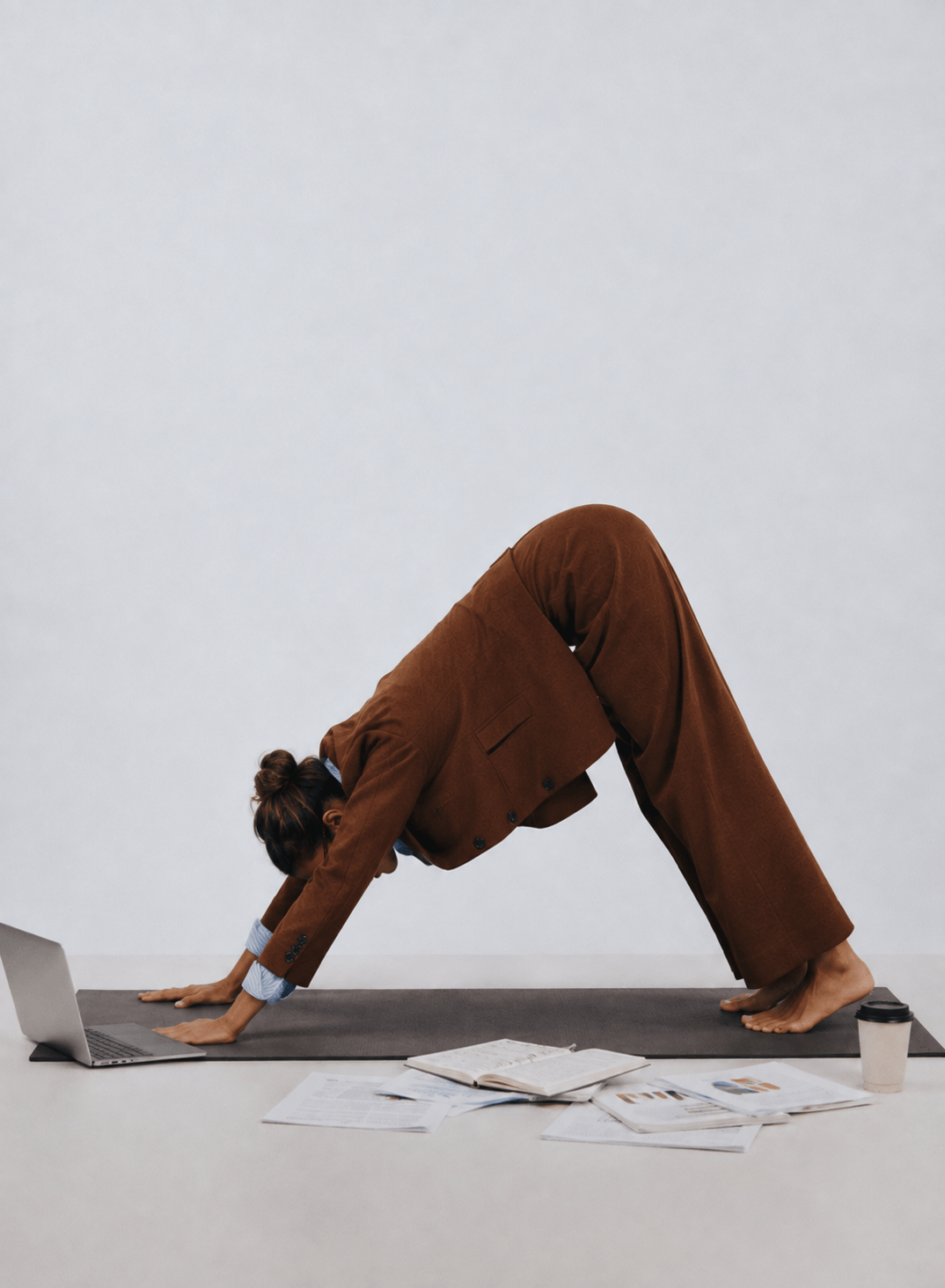 Woman with brown suit practicing yoga in a forward bend pose on a mat in front of a laptop and scattered papers, with a coffee cup beside her.