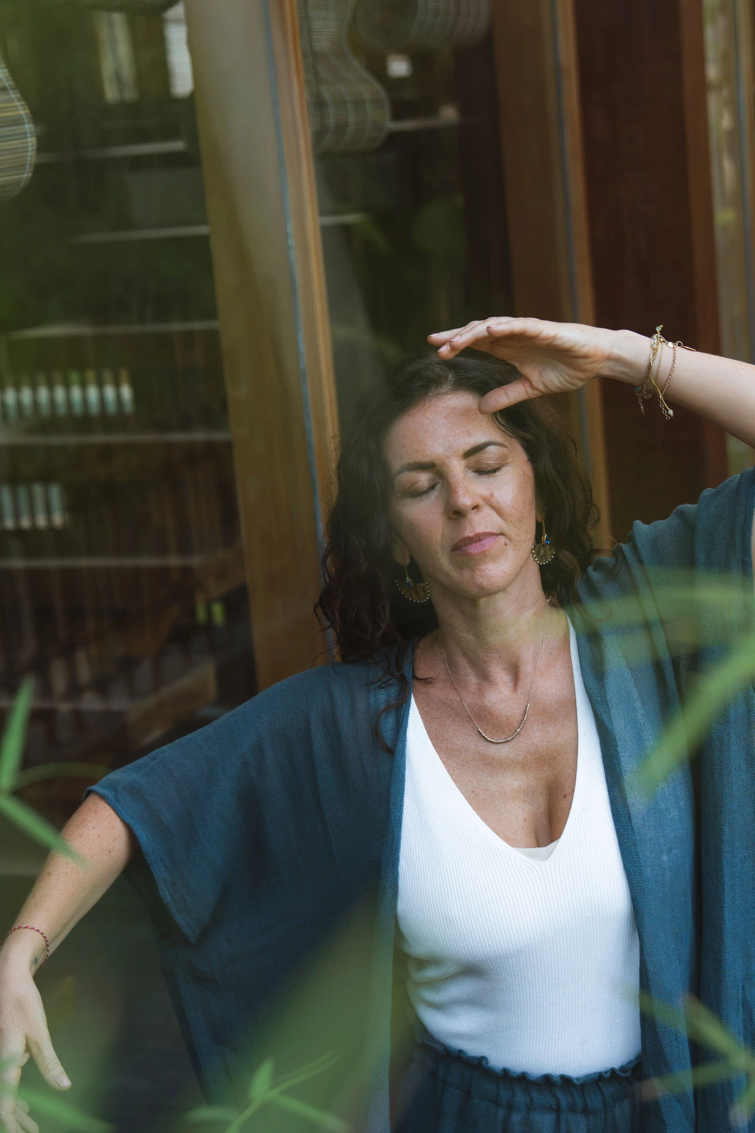 A woman with dark curly hair and light skin is closing her eyes and holding her hand above her forehead. She is dressed in a white top and a blue cardigan, and is wearing jewelry including a necklace and bracelets. The photo appears to be taken through a glass window with some greenery visible in the foreground, and a wooden structure in the background.