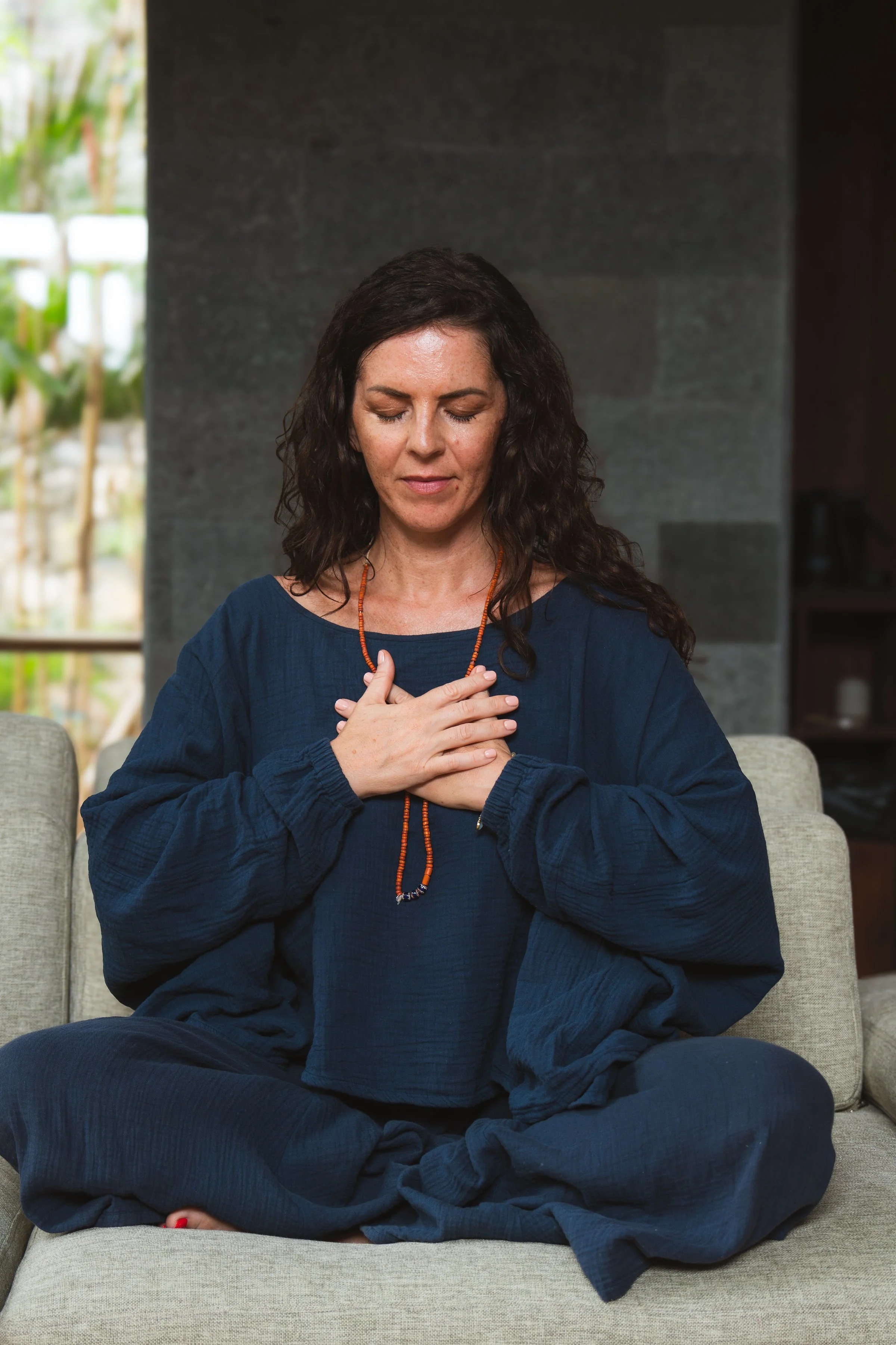 A woman sitting cross-legged on a beige couch with eyes closed, hands over her chest, in a peaceful meditation pose, wearing a navy blue long-sleeve top and a beaded necklace.