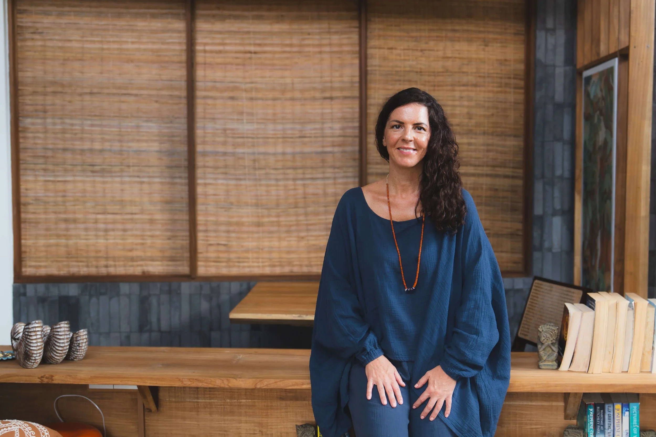 A woman with long dark curly hair wearing a blue dress and a beaded necklace standing indoors in front of a wooden and bamboo wall with books and decorative items on a table.