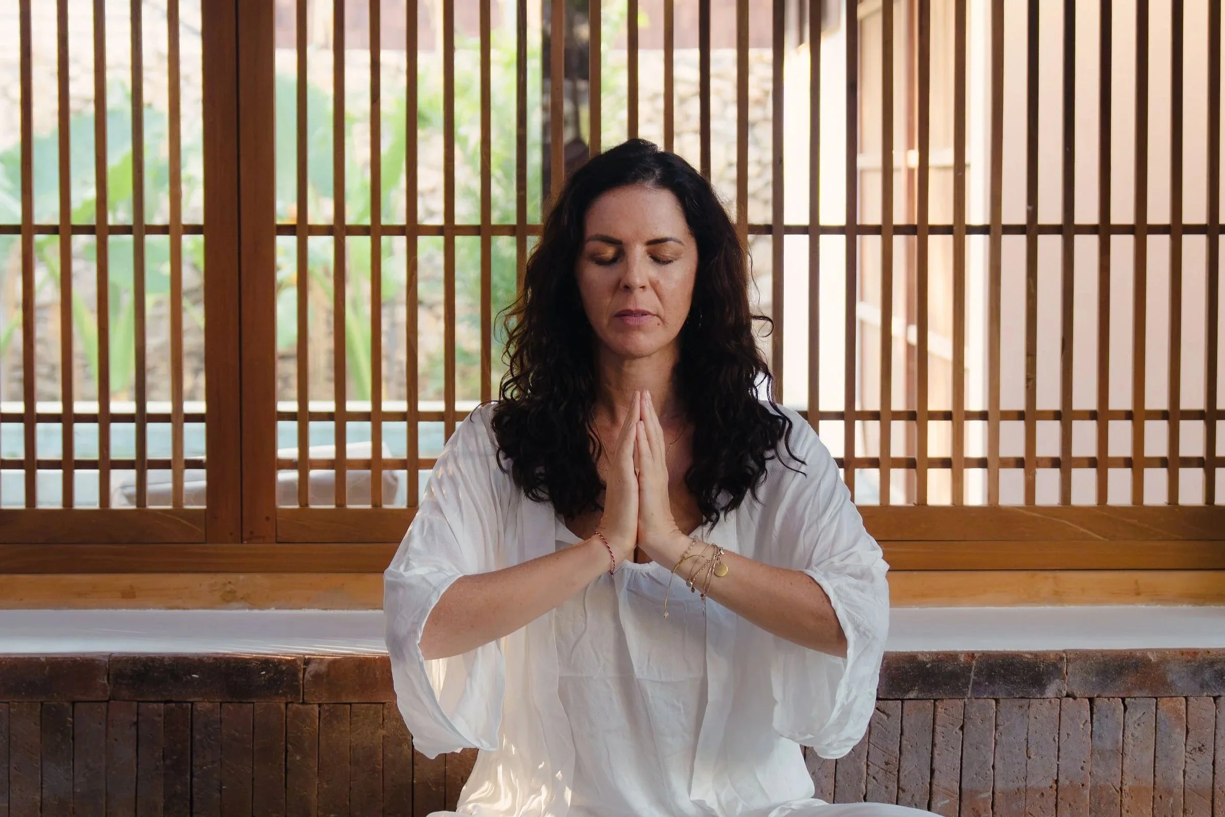 A woman with dark, curly hair, wearing a white top, sitting cross-legged with hands pressed together in prayer or meditation, eyes closed, in front of a wooden grid screen with a natural outdoor background.