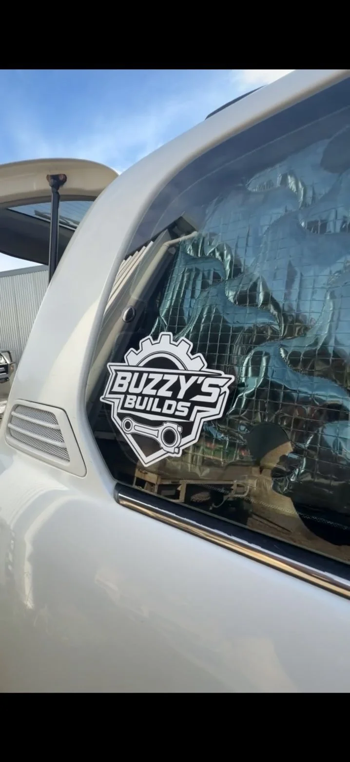 Close-up of a vehicle window with a Buzzy's Builds sticker, revealing a reflecting construction or building project inside.