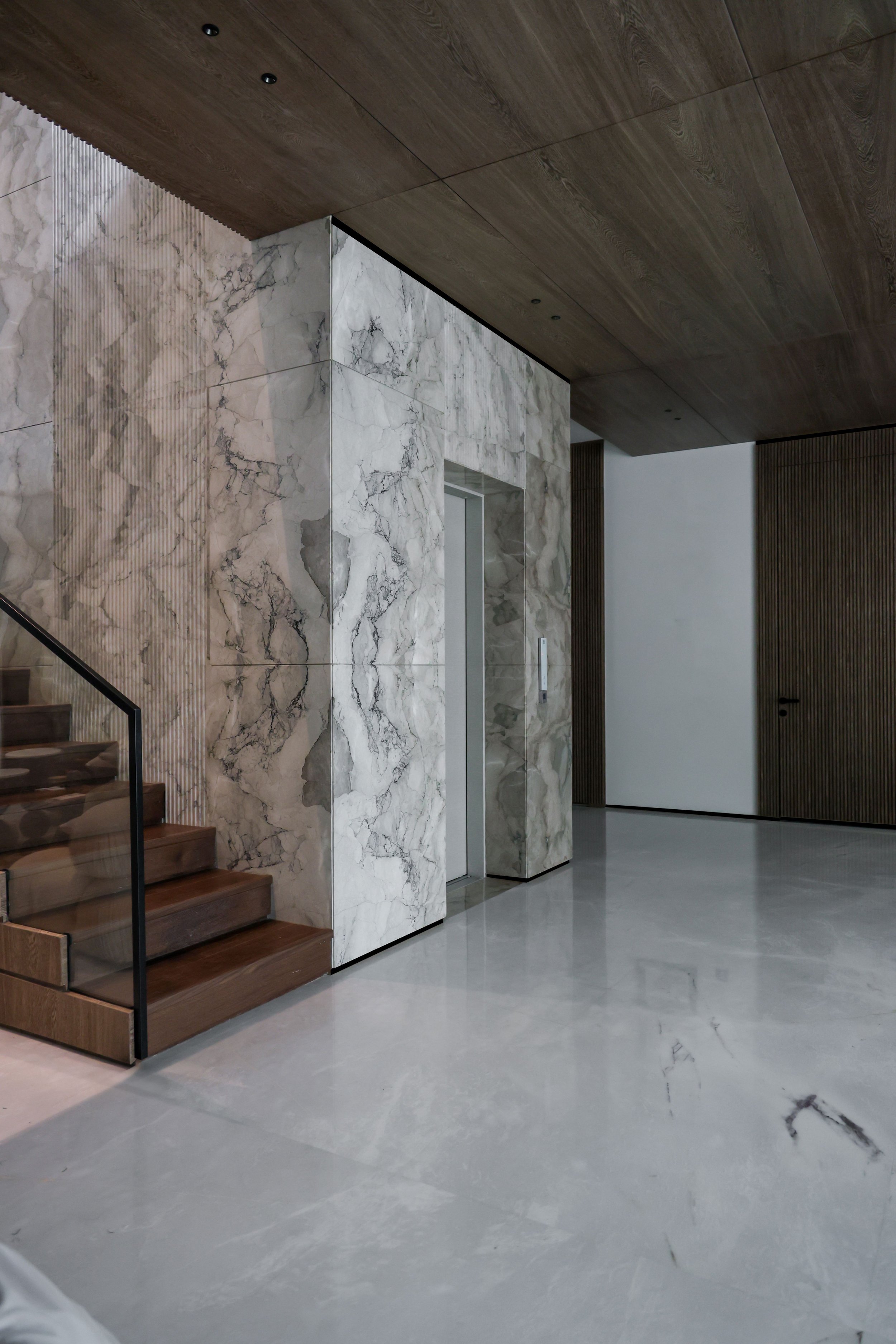 Interior modern lobby with marble flooring, marble walls, wooden ceiling, and stairs with glass railing.
